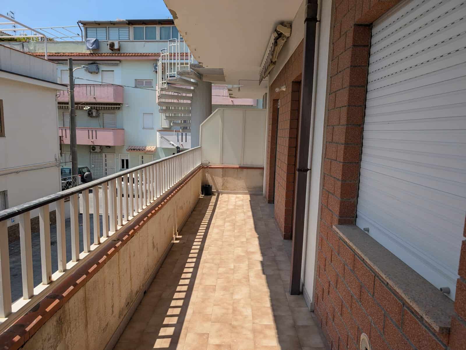 Balcone di un appartamento in zona urbana con ringhiera in metallo e pavimentazione in piastrelle, vista su edifici residenziali e scale esterne.