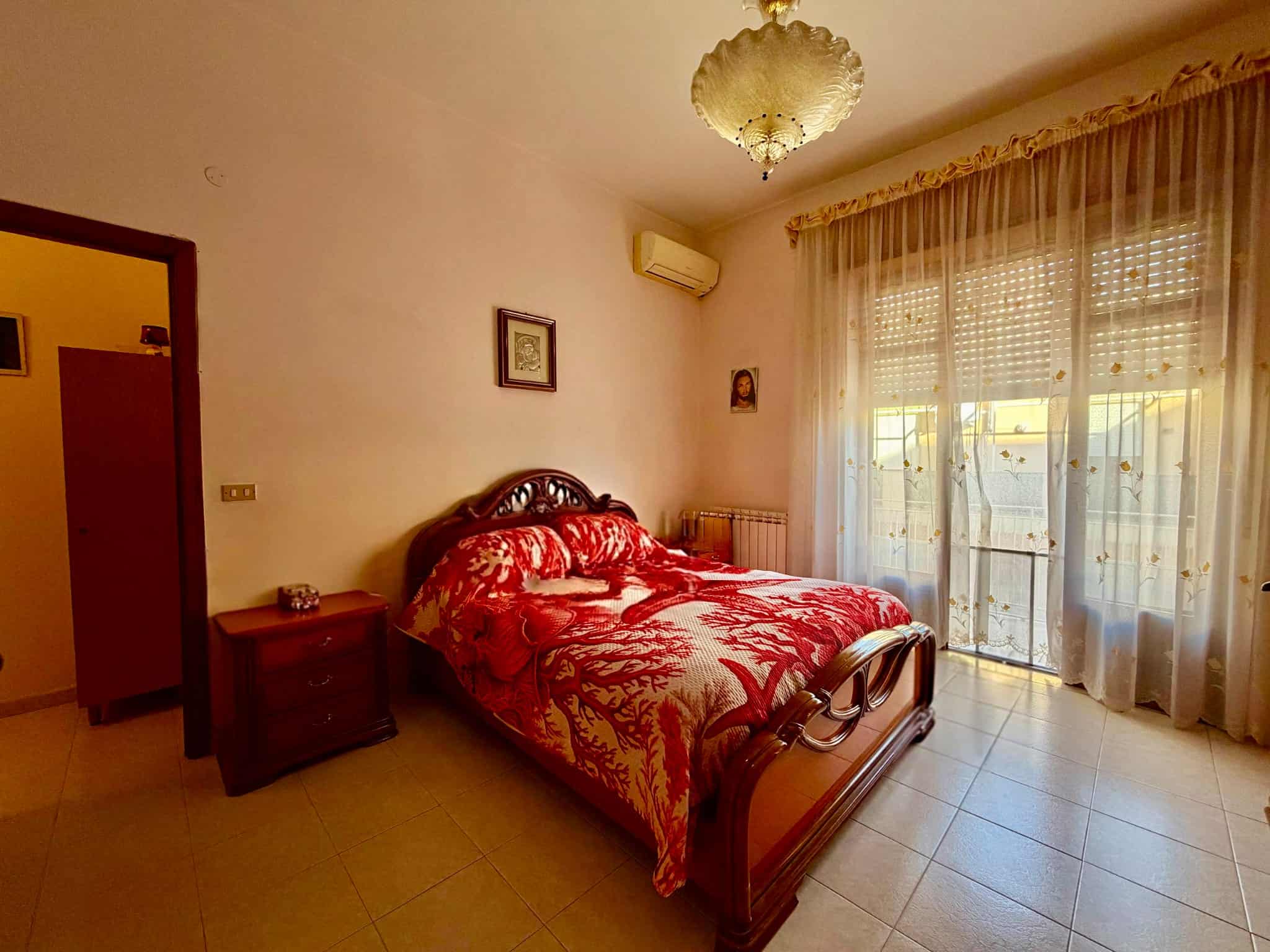 Camera da letto luminosa con letto in legno, copriletto rosso e tende trasparenti che lasciano entrare molta luce naturale.
