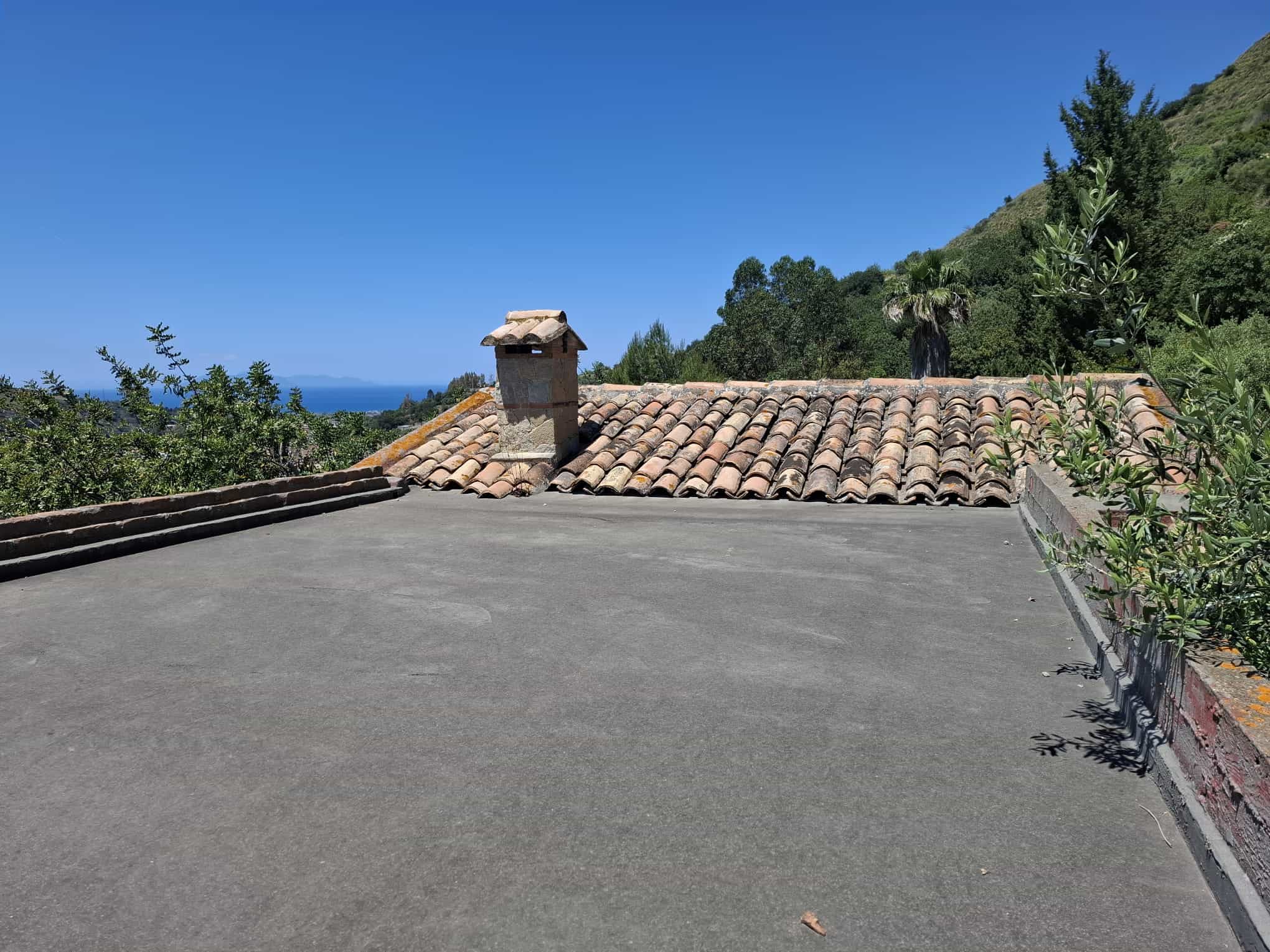 Il tetto di una casa con tegole in terracotta, circondato da natura e colline verdi, sotto un cielo azzurro, in un paesaggio tranquillo e panoramico.