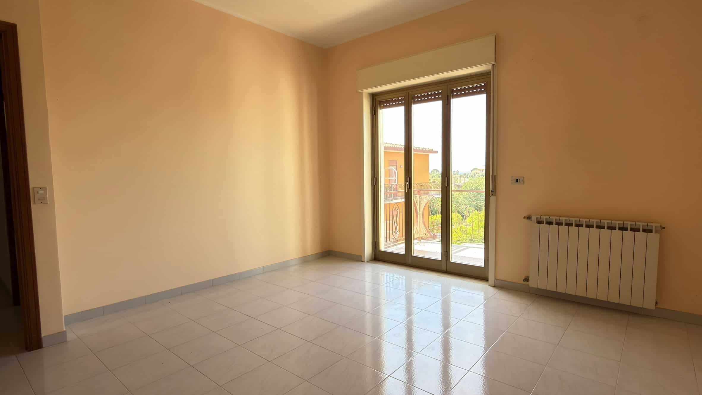 Stanza vuota con pareti beige, grande finestra con vista su balcone e radiatore, pavimento in piastrelle chiare, ambiente luminoso e spazioso.