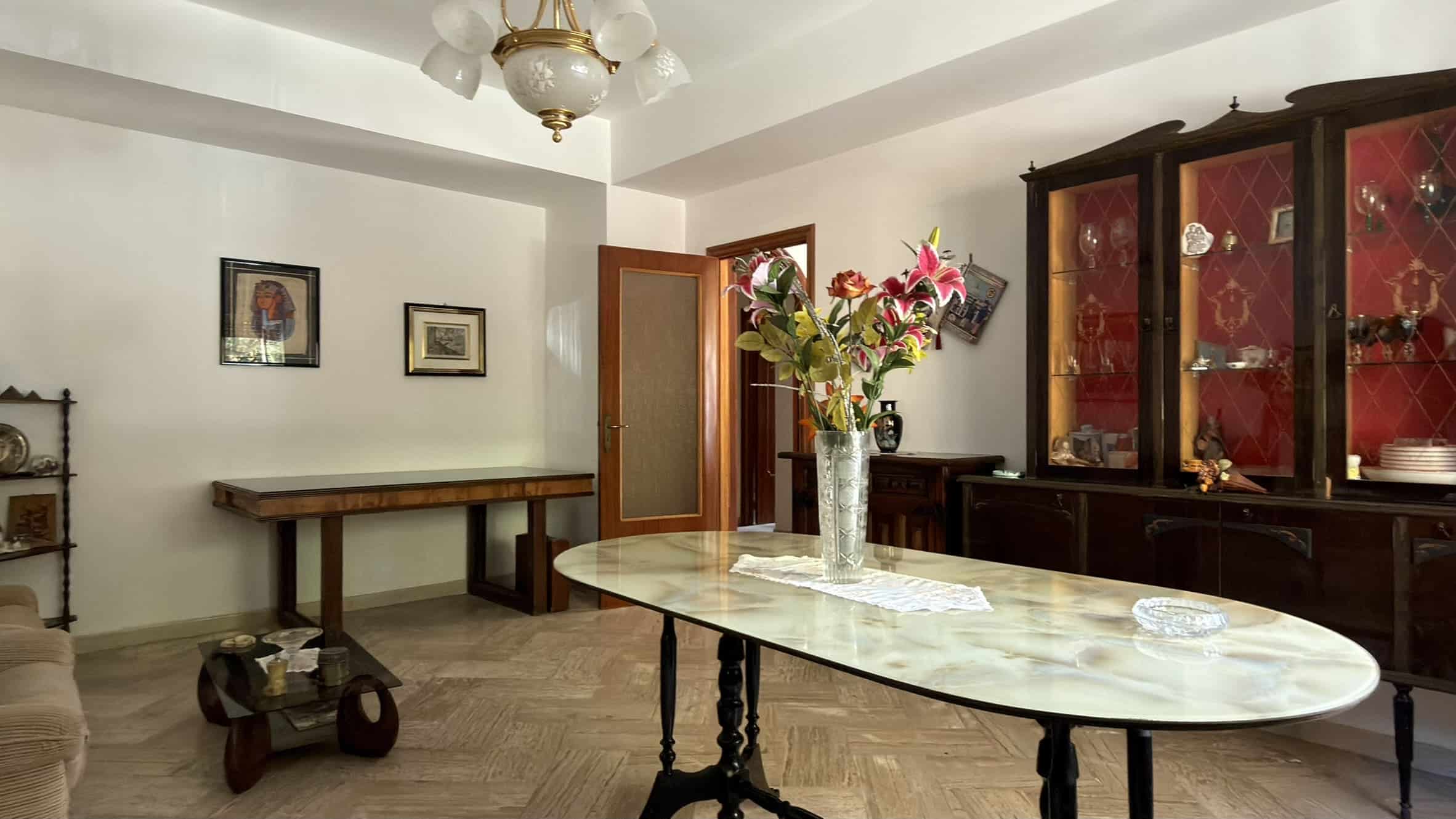 Interno di una sala da pranzo con tavolo in marmo, vetrina in legno e decorazioni floreali, ambiente luminoso e accogliente.