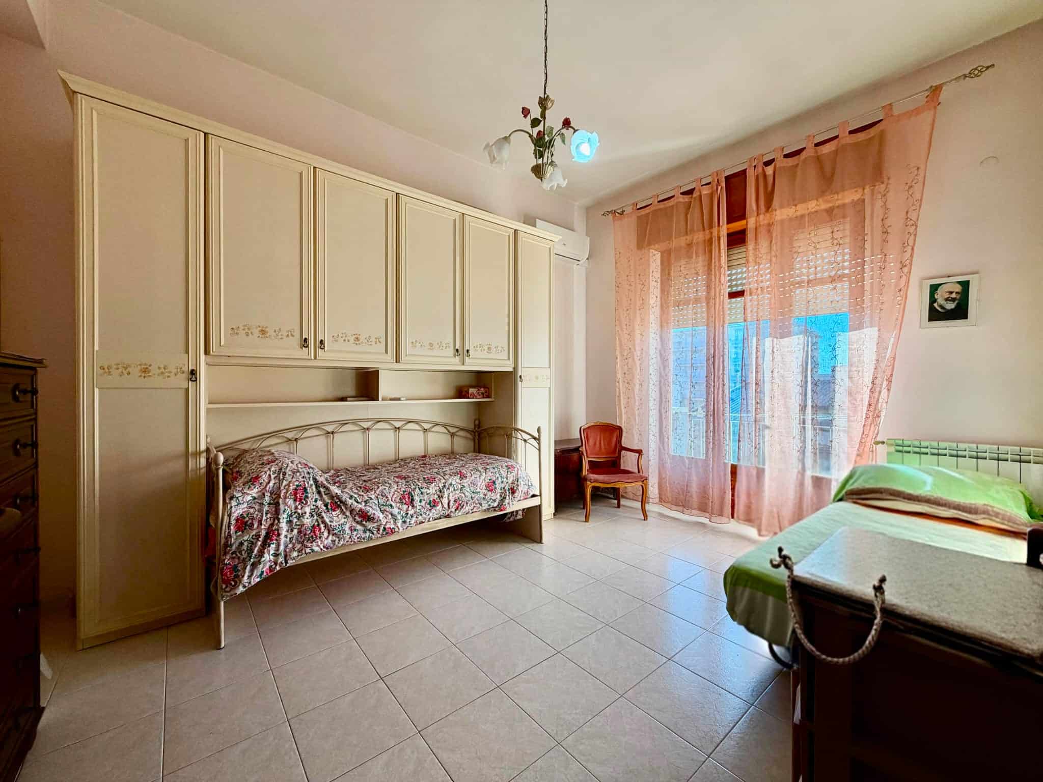 Camera da letto spaziosa con armadio a muro, letto singolo, finestra con tende rosa e arancioni, e mobili classici in stile vintage.