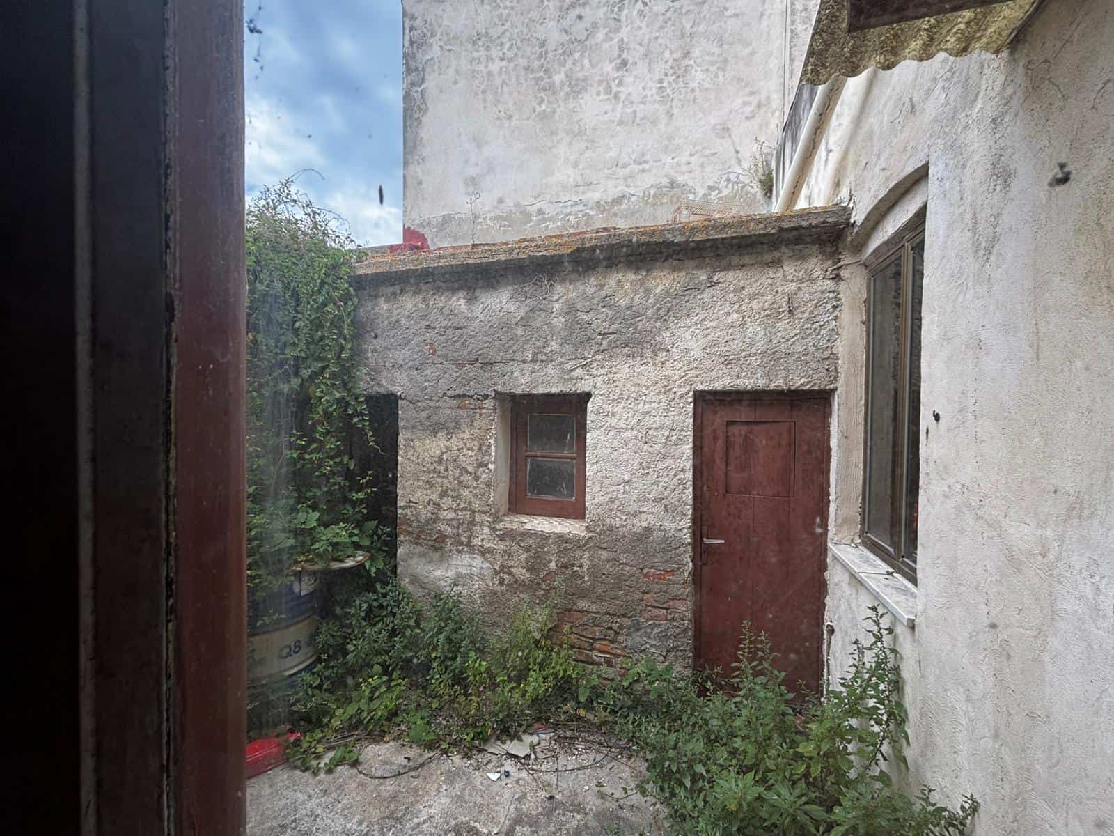 Vecchio cortile esterno con pareti in pietra e legno, piante selvatiche e porta in legno rovinata, ambiente rustico e abbandonato, ideale per ristrutturazione o progetto di recupero immobiliare.