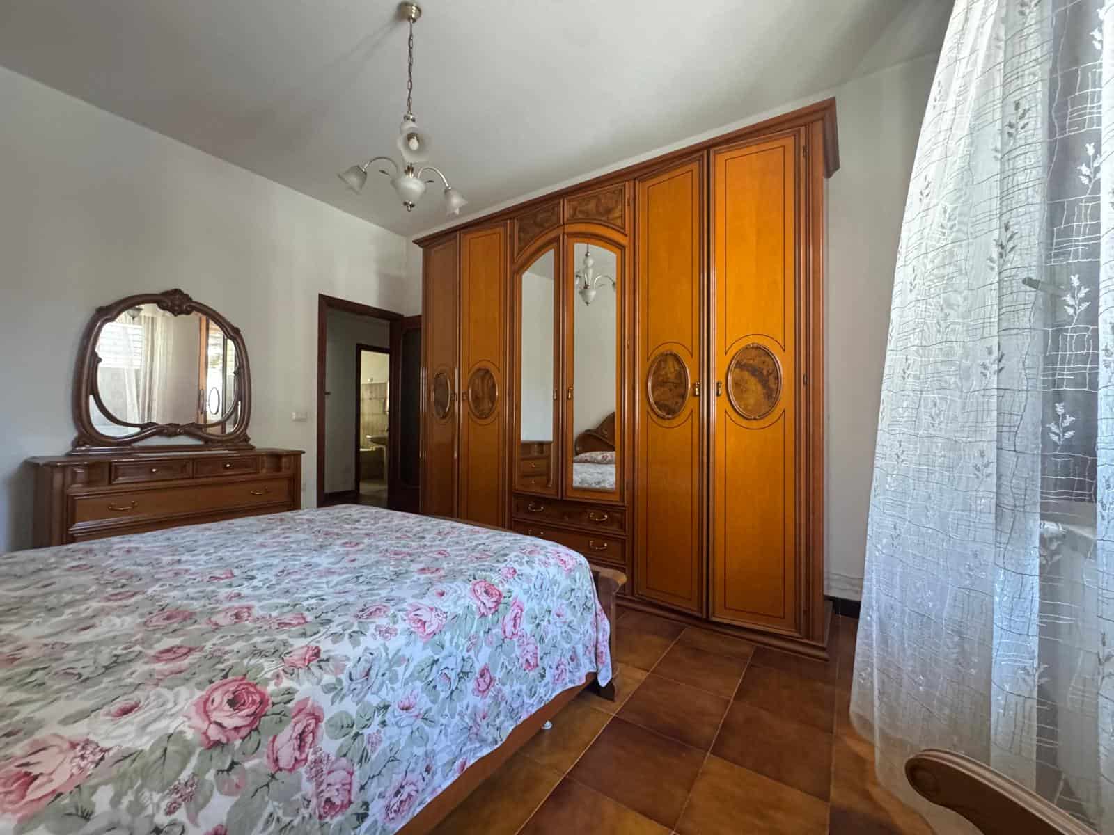 Il letto matrimoniale con copriletto floreale in una camera da letto classica, con grande armadio in legno e specchio, e una finestra con tende leggere, in un ambiente luminoso e accogliente.