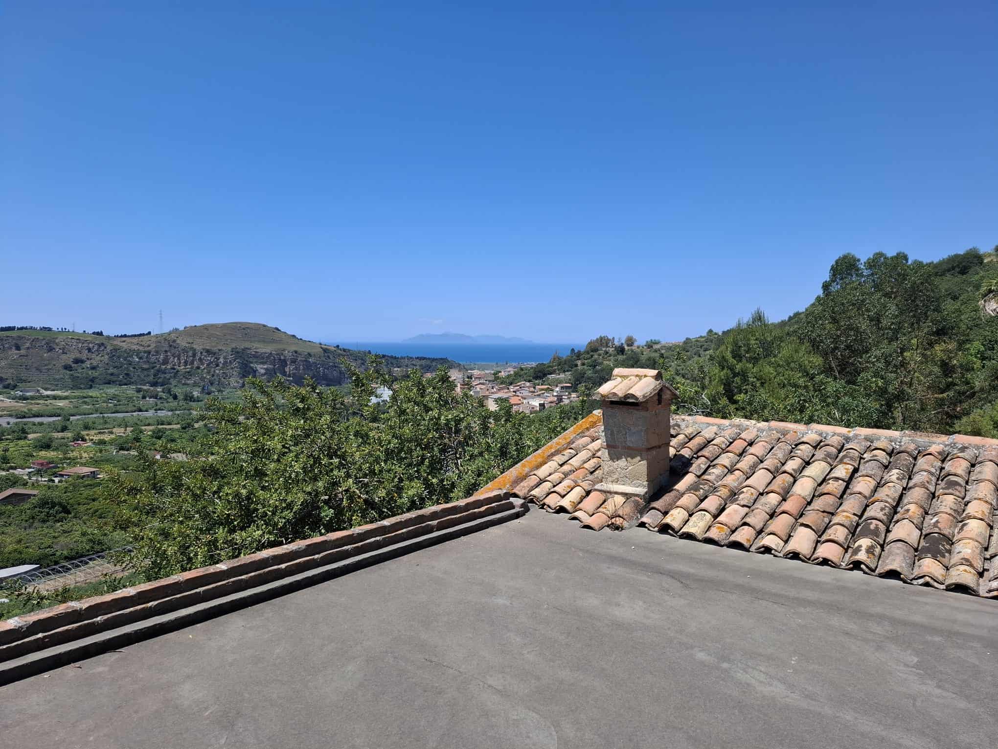 Vista panoramica dal tetto di una casa in campagna con paesaggio collinare e mare sullo sfondo, cielo sereno, ideale per immobili in vendita o affitto in zone rurali o costiere.