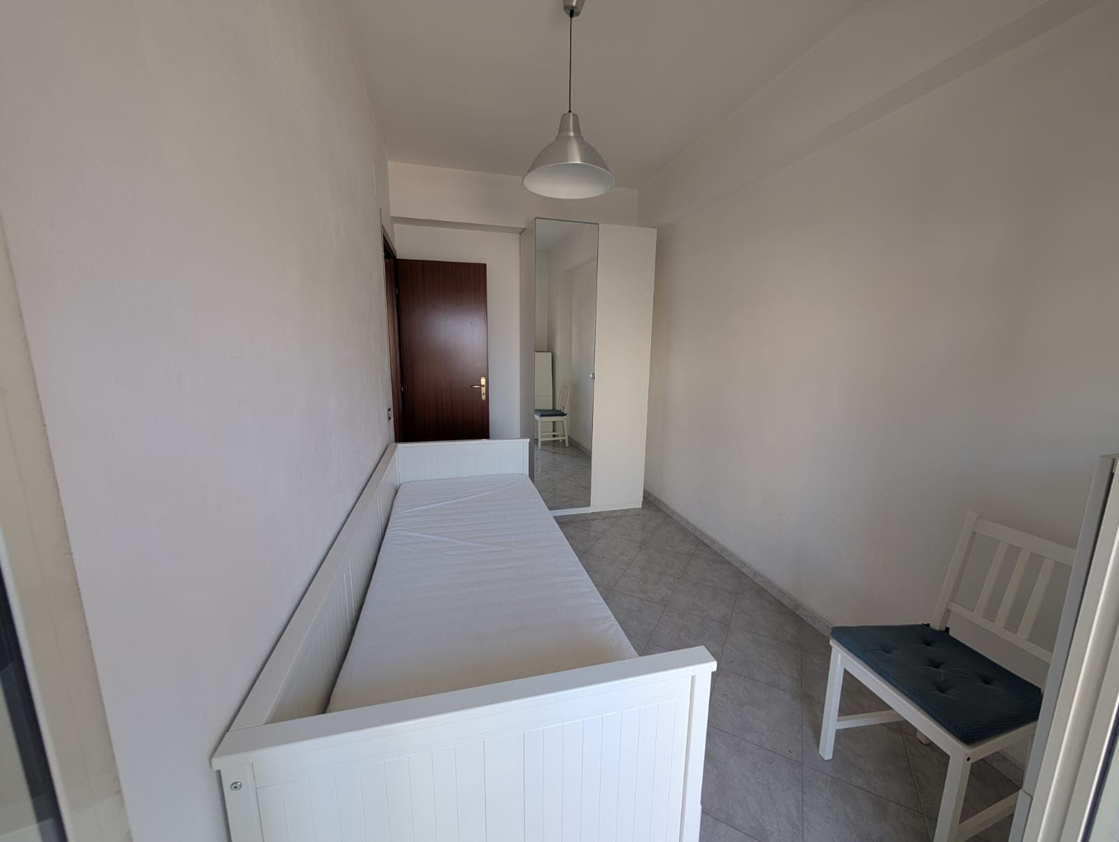 Camera singola arredata con letto e sedia, ambiente luminoso e semplice.