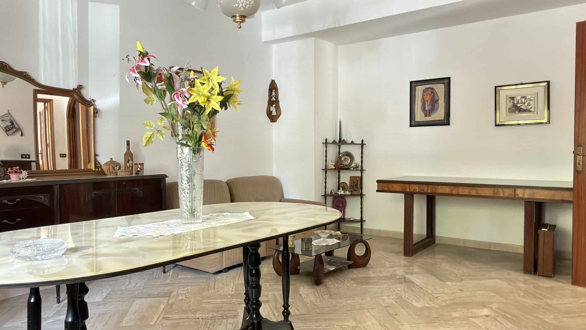 Interno di una casa con tavolo, sedie, e decorazioni vintage, ambiente luminoso e accogliente.