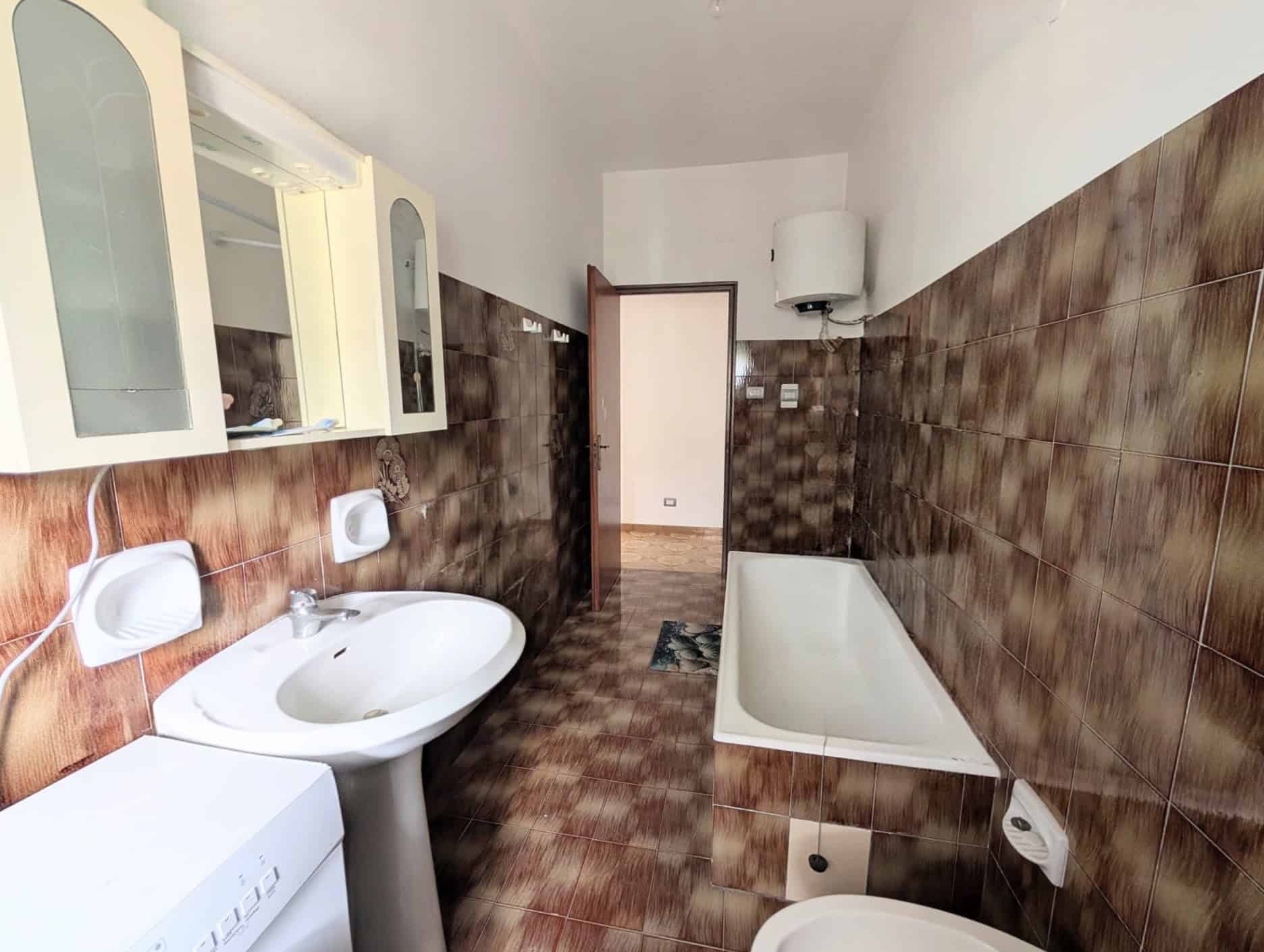 Bagno con vasca, lavabo e armadietto a muro, rivestimenti in piastrelle marroni, ambiente semplice e funzionale.