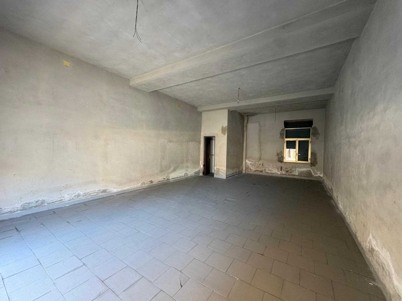 Interno di un appartamento in fase di ristrutturazione con pareti e soffitto in cemento grezzo e pavimento in piastrelle.