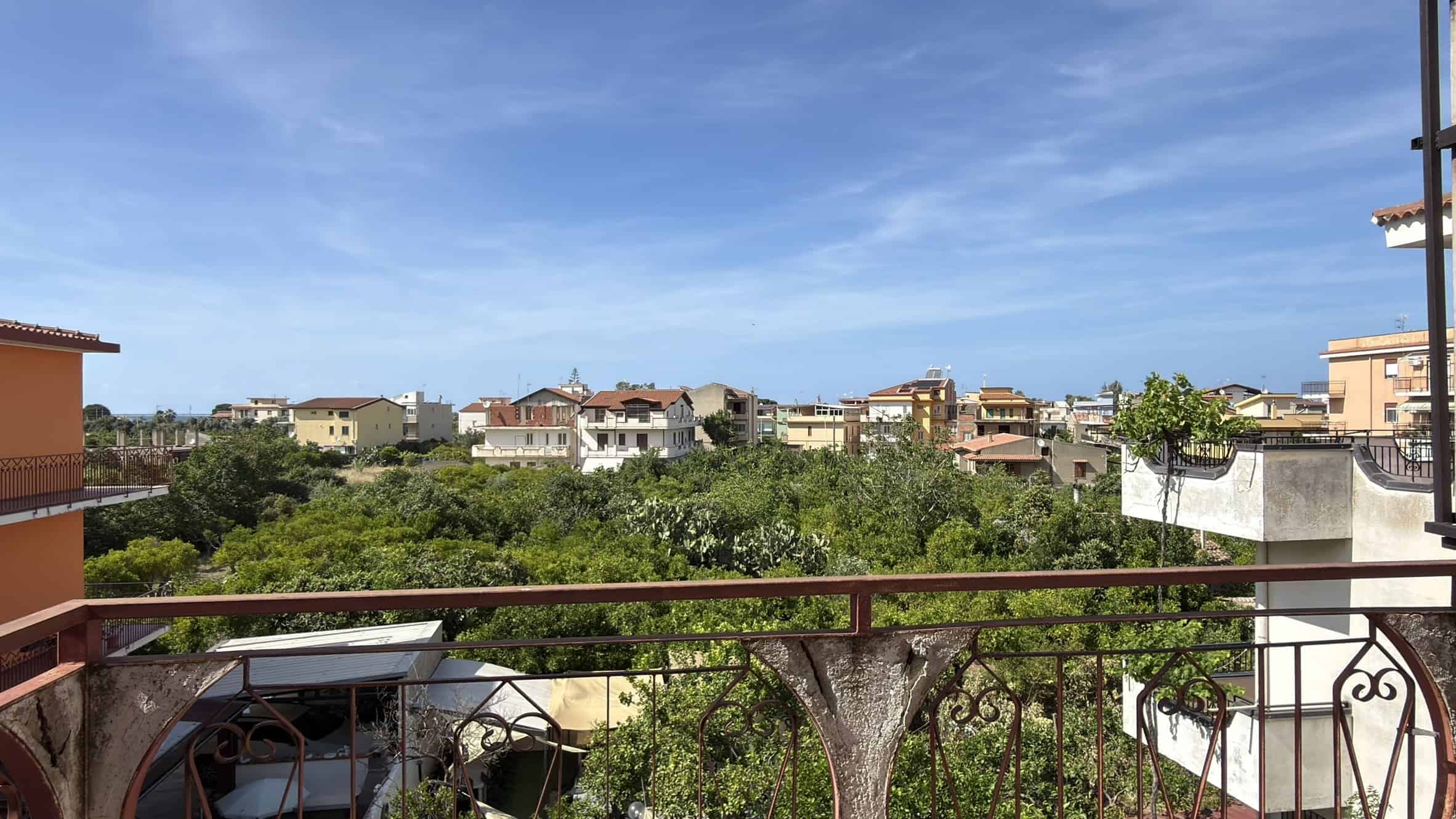 Vista panoramica di un quartiere residenziale con case e alberi sotto un cielo sereno, vista da un balcone con ringhiera in ferro battuto.