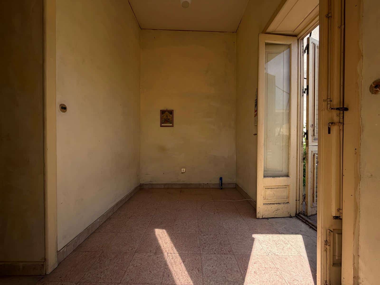 Interno di una stanza vuota con pareti beige, pavimento in piastrelle e balcone con porta finestra aperta, luce naturale.
