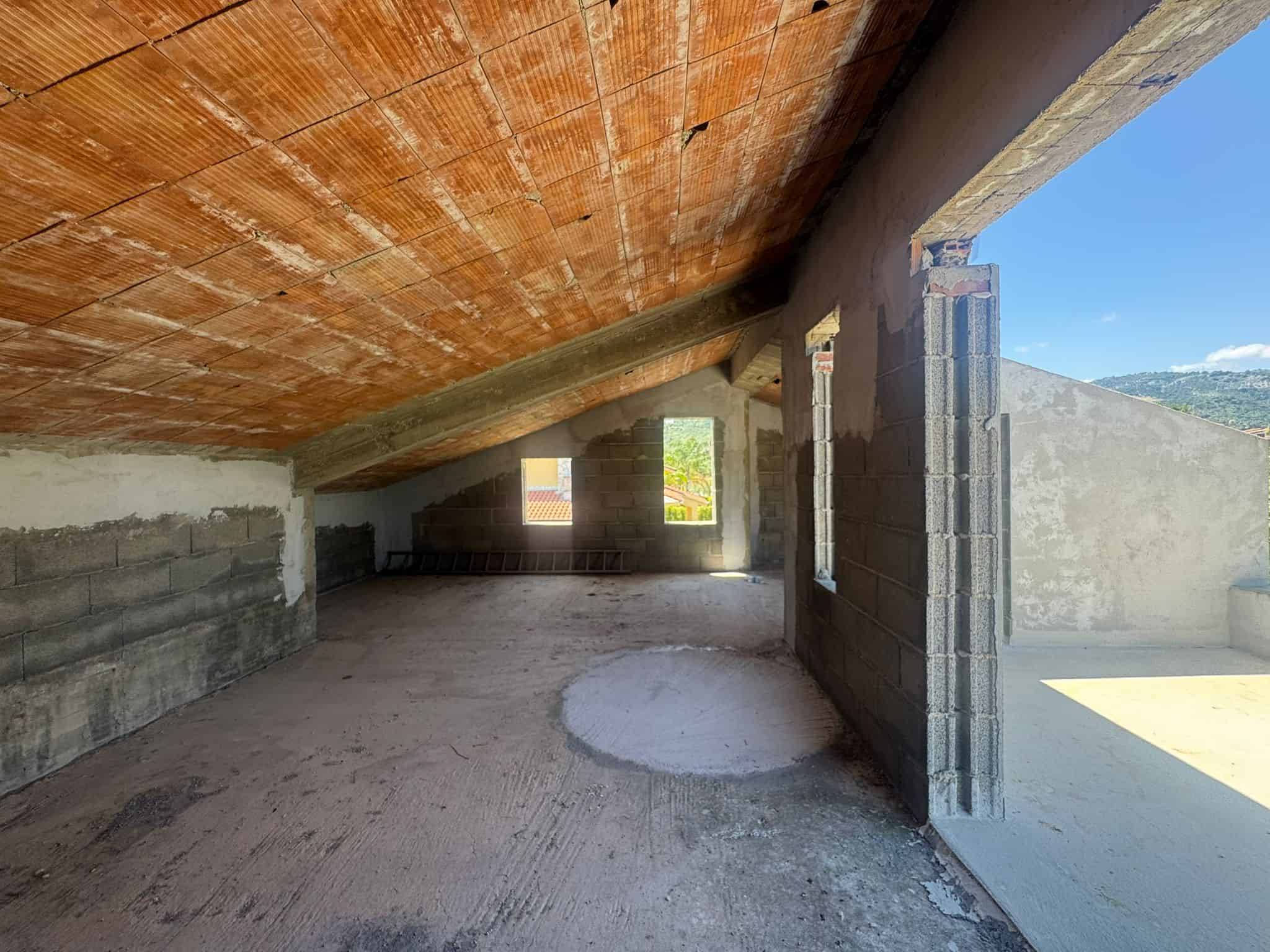 Interno di un edificio in costruzione con soffitto in cemento e pareti in mattoni, vista verso l'esterno con cielo azzurro e paesaggio montano.