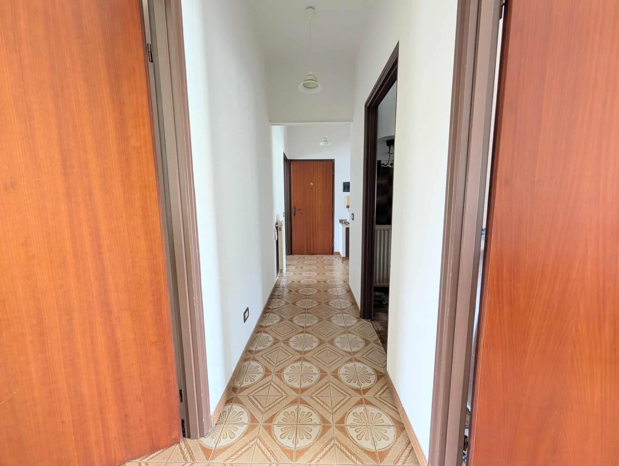 Ingresso di un appartamento con pavimento in piastrelle decorative e pareti bianche, vista verso la porta d'ingresso in legno.
