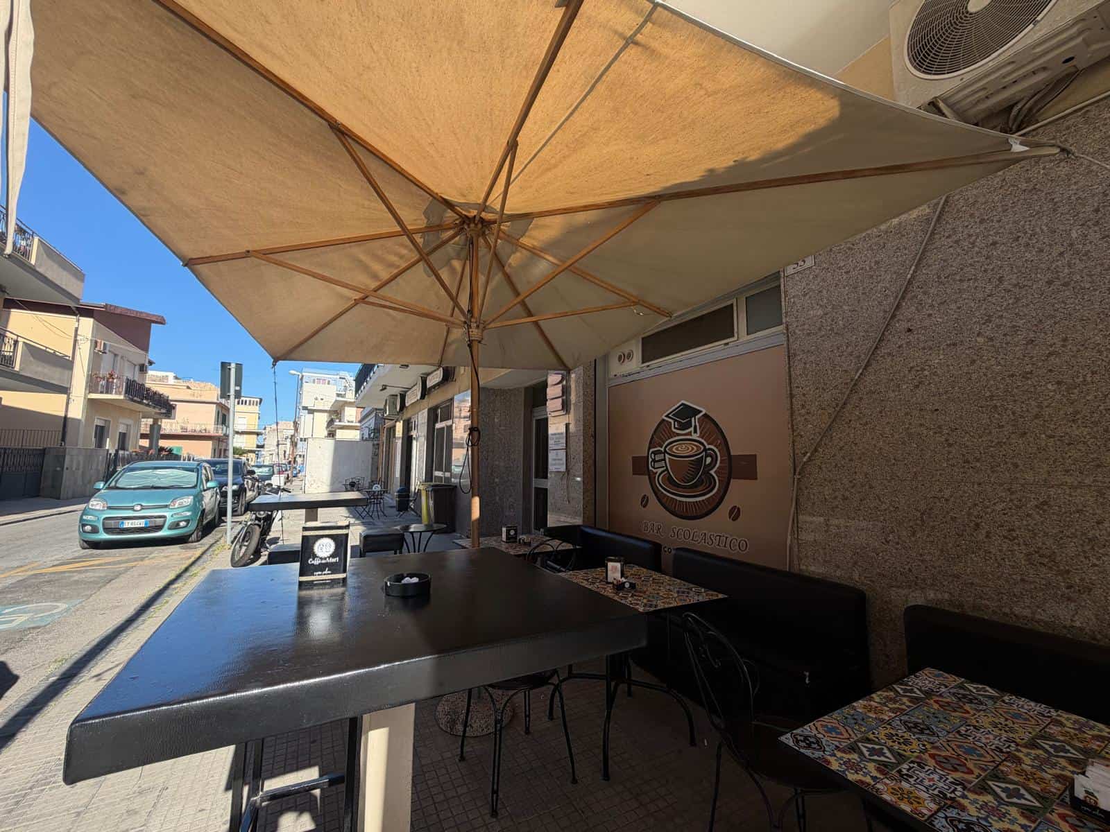 Spazio esterno di un'attività commerciale con tavoli, sedie e grande ombrellone, situato in Via Risorgimento 86, Milazzo, ME.