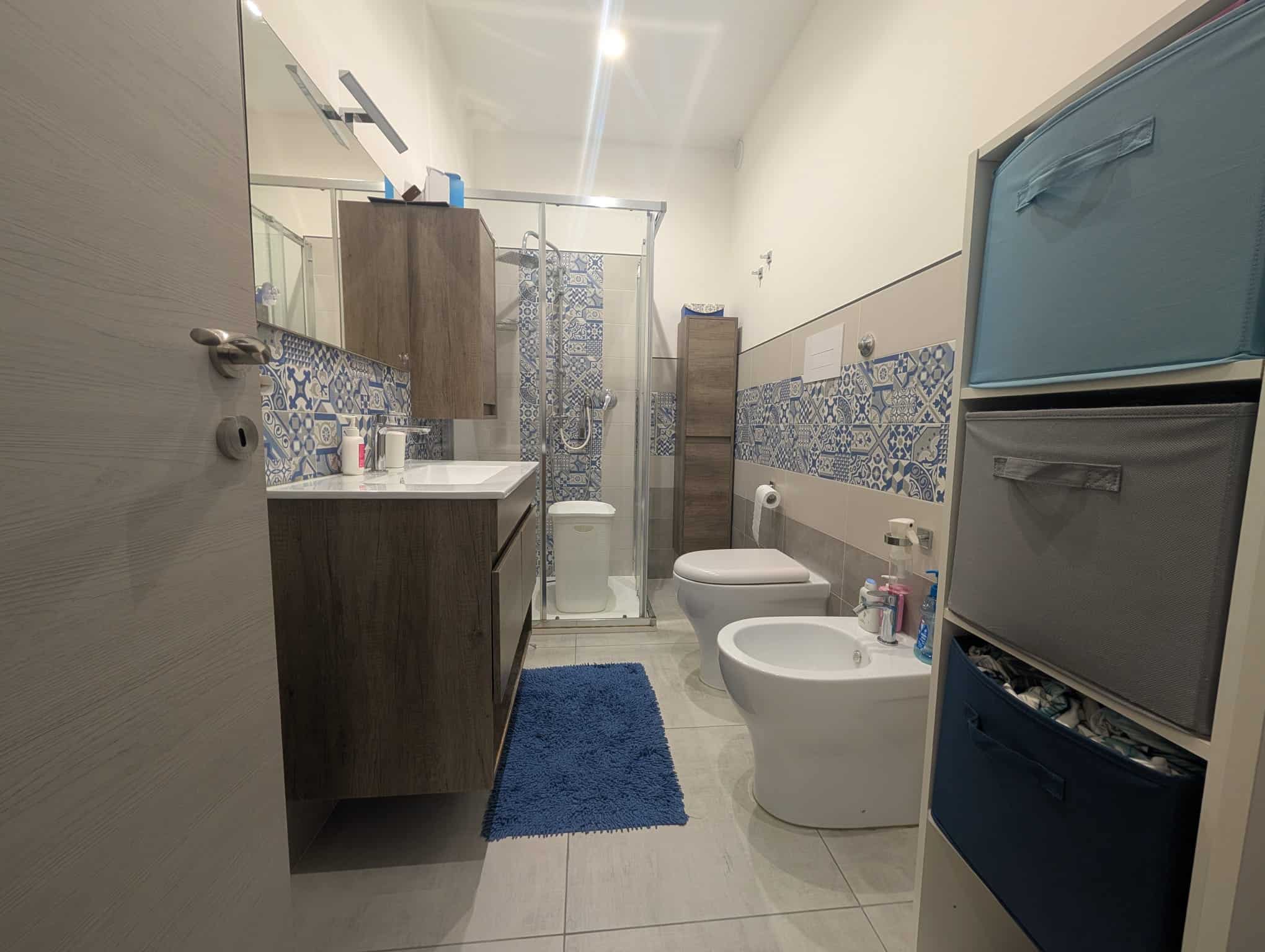 Lavabo e servizi igienici moderni in bagno spazioso con piastrelle decorative blu e bianco, armadietti in legno, doccia con porta in vetro e accessori essenziali per il comfort quotidiano.