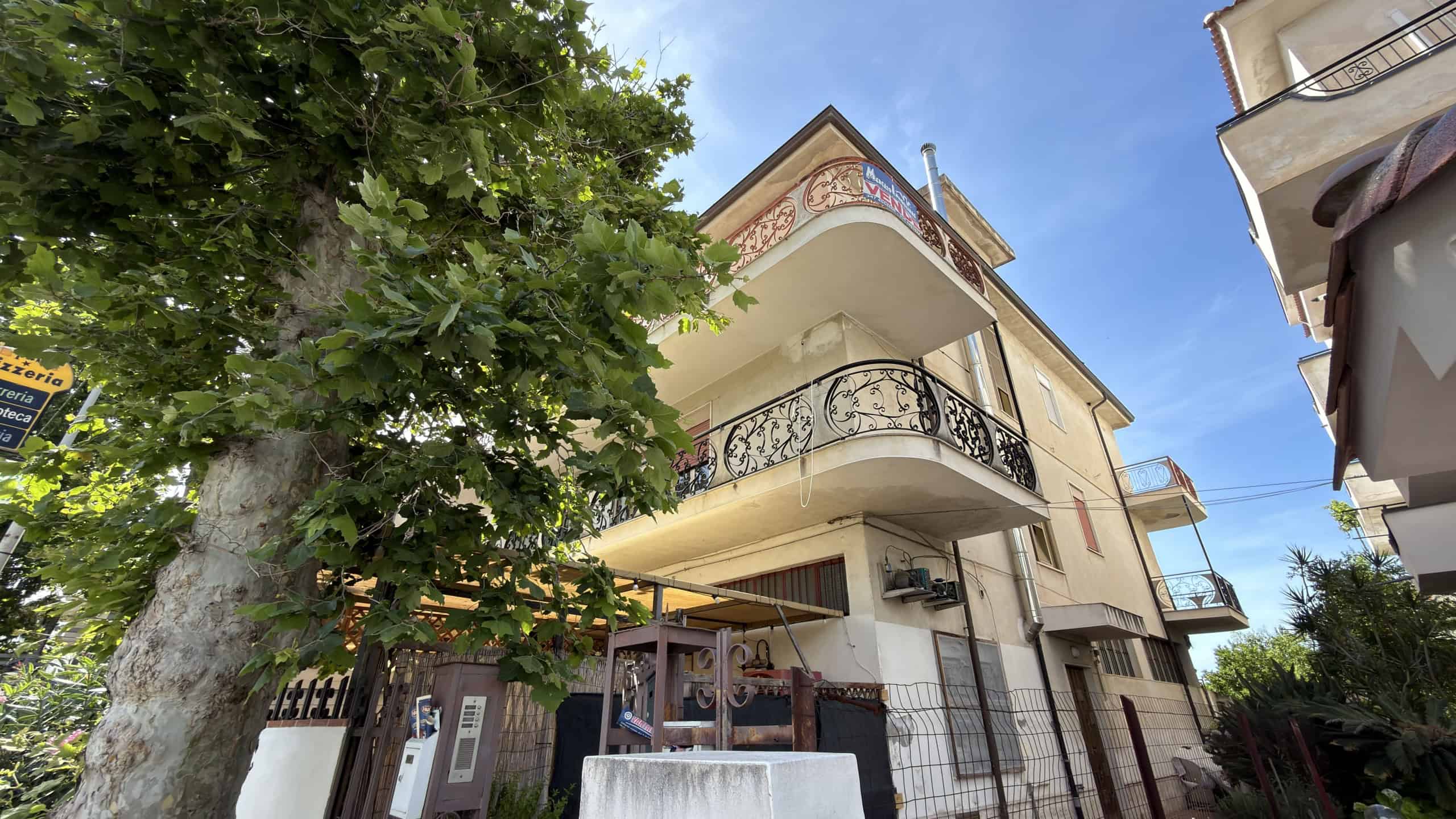 Immobile residenziale a tre piani con balconi e terrazze, circondato da alberi e con un cielo sereno sullo sfondo.