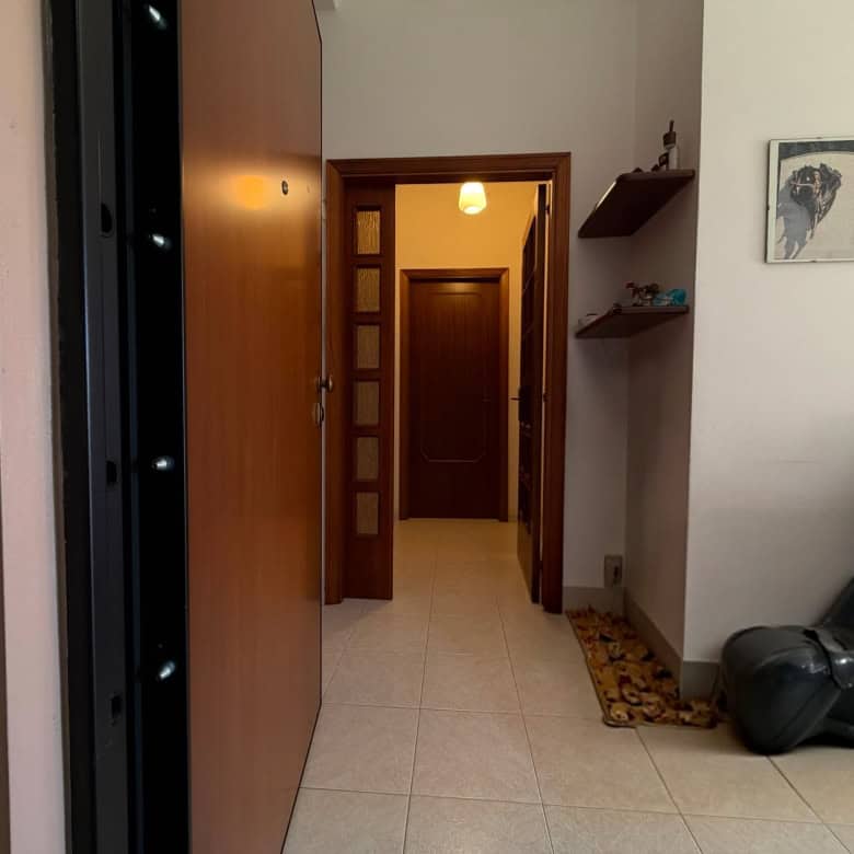 Ingresso di un appartamento con porta in legno e corridoio ben illuminato, arredato con scaffali e decorazioni.
