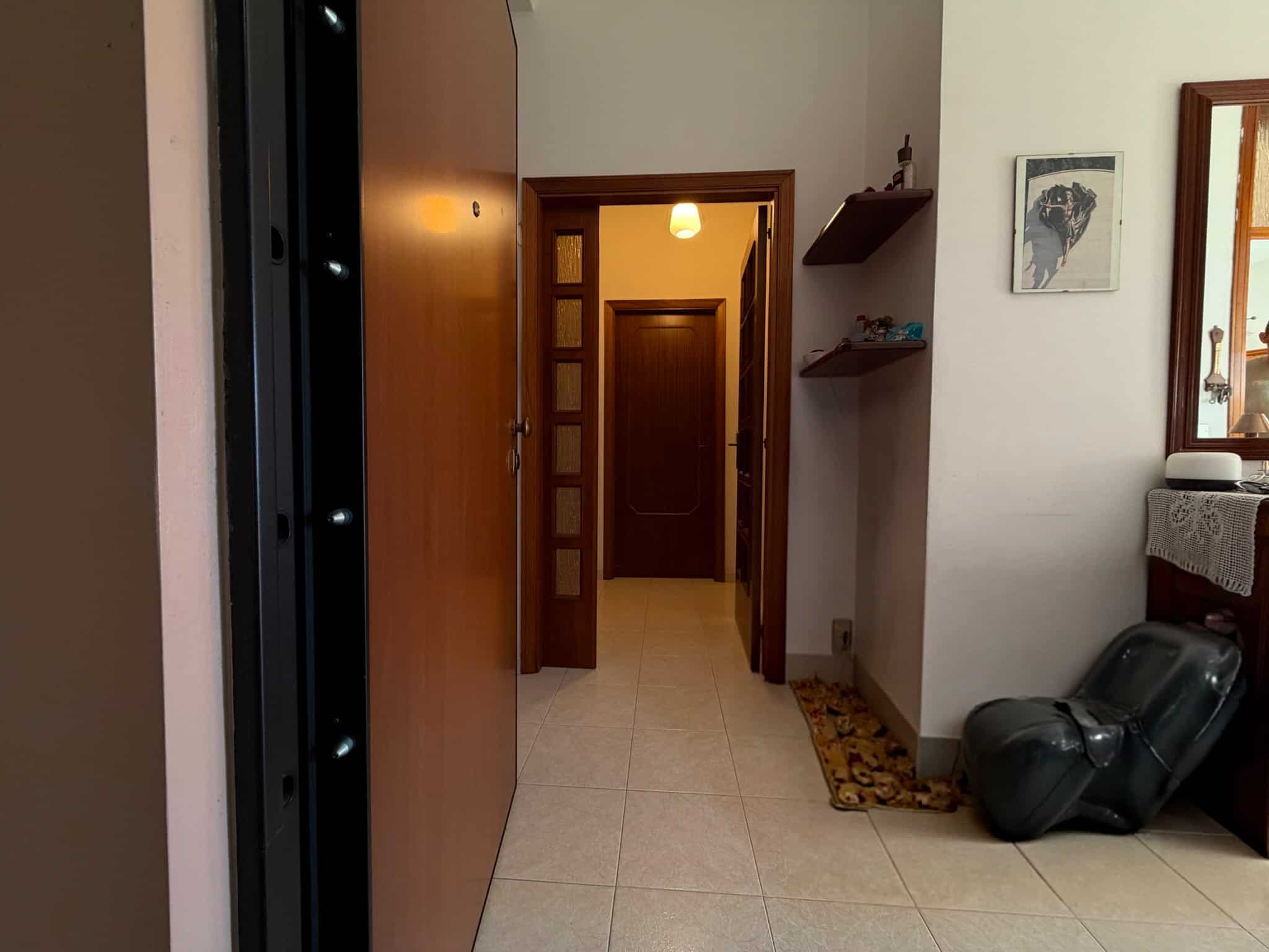 Ingresso di un appartamento con porta in legno e corridoio ben illuminato, arredato con scaffali e decorazioni.
