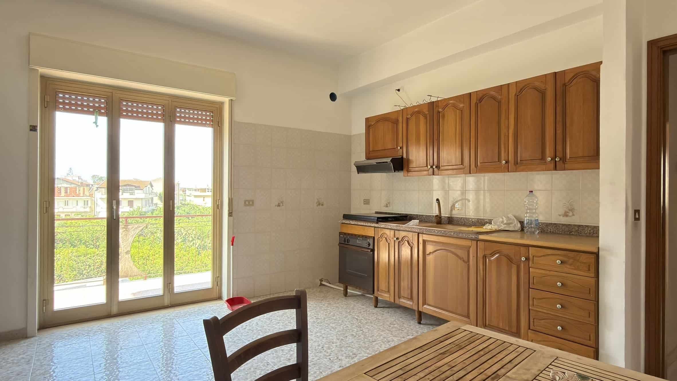 Cucina spaziosa con mobili in legno e accesso a balcone con vista sul verde e le case circostanti.