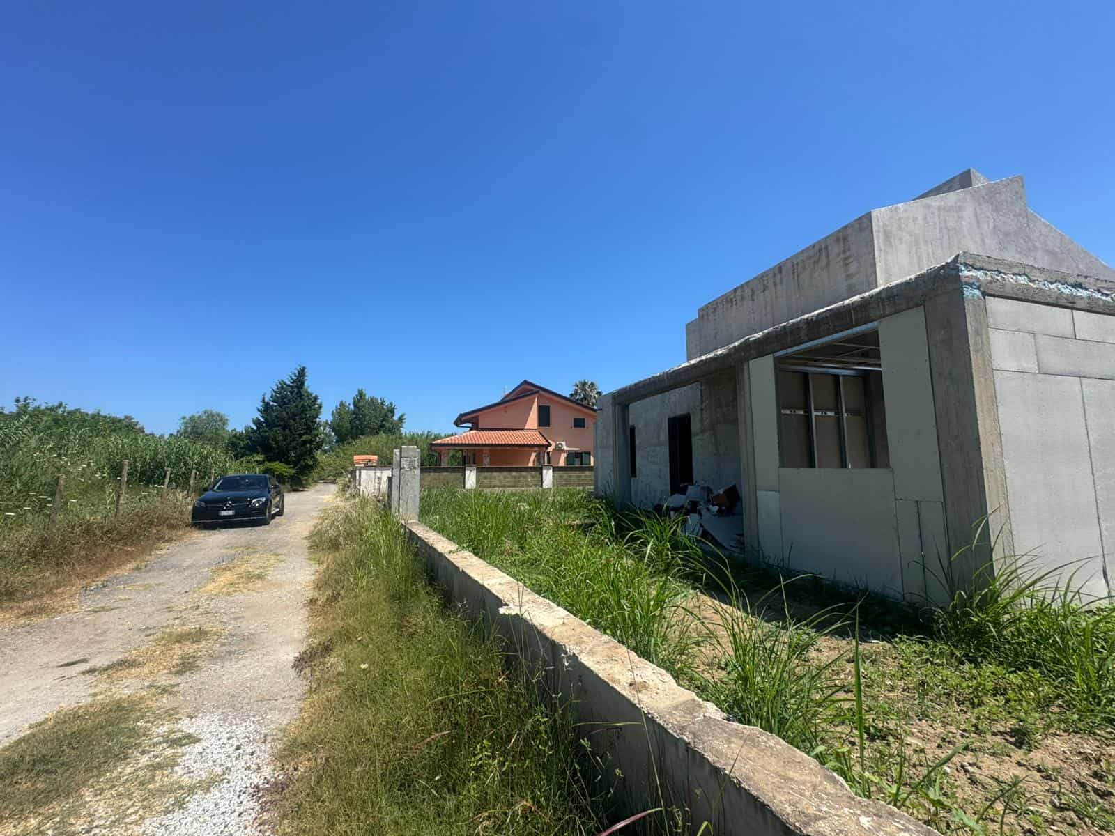 Cantiere edile con edificio in costruzione e strada sterrata in campagna sotto un cielo azzurro.