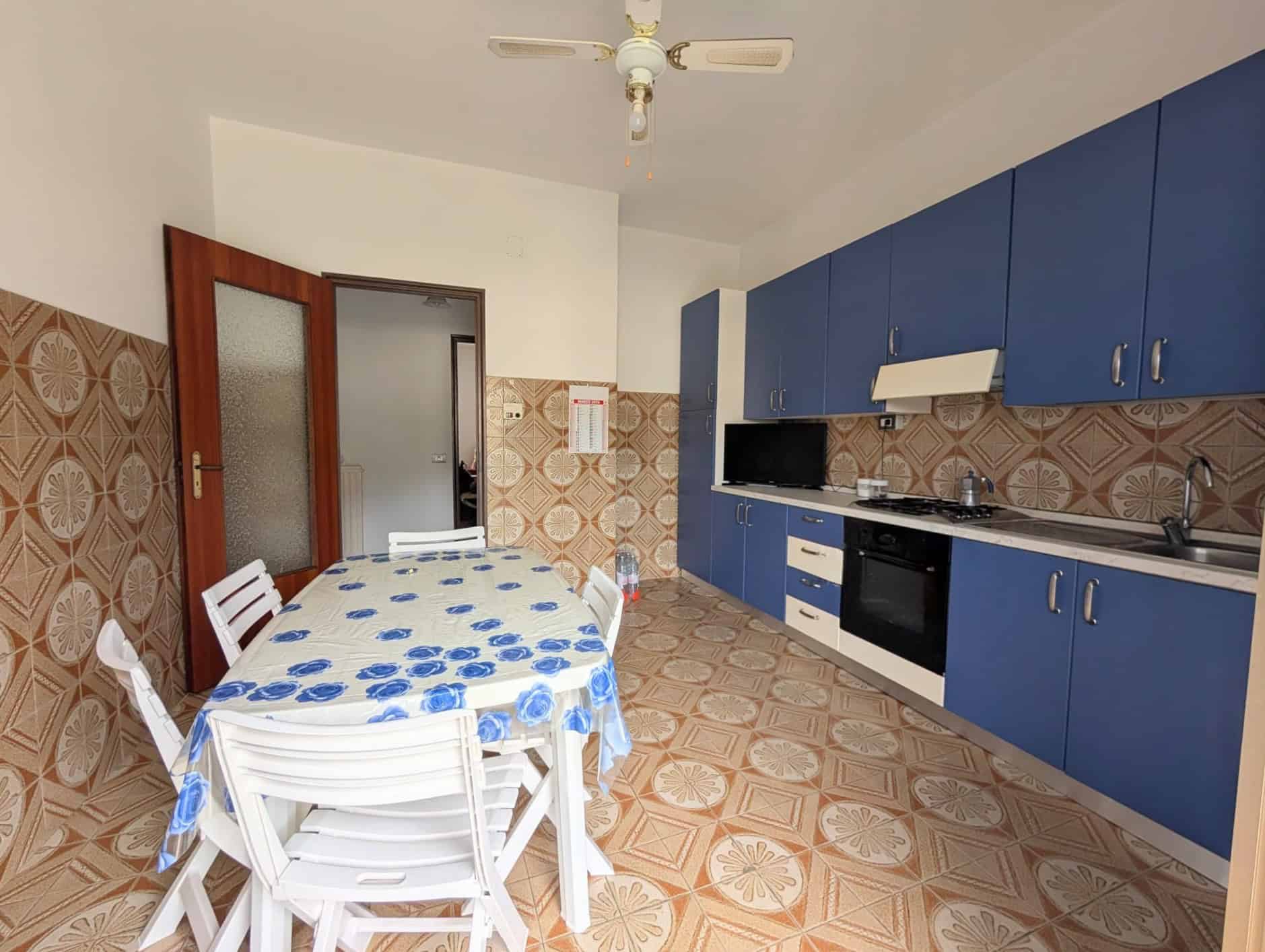 Cucina spaziosa con mobili blu, tavolo rotondo con tovaglia a fiori blu e sedie bianche, pareti rivestite con piastrelle decorative.