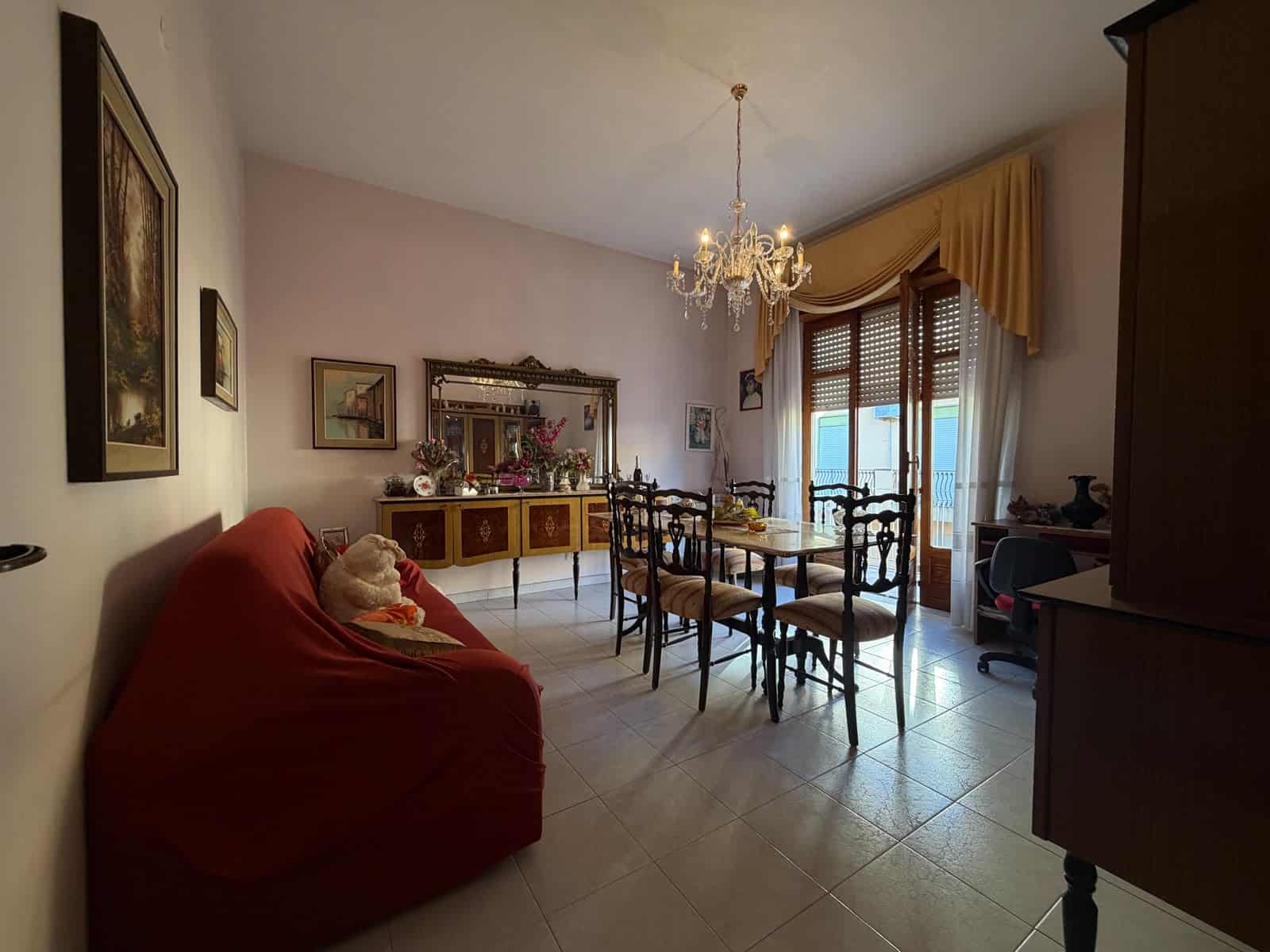 Interno di un appartamento con soggiorno arredato, tavolo, sedie, divano rosso e lampadario elegante.