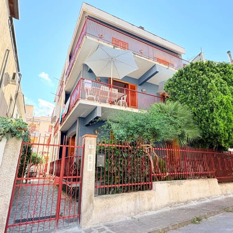 Balcone con ombrellone e piante verdi in un edificio residenziale a più piani, con recinzione rossa e accesso pedonale, in un quartiere urbano soleggiato e tranquillo, in Italia.
