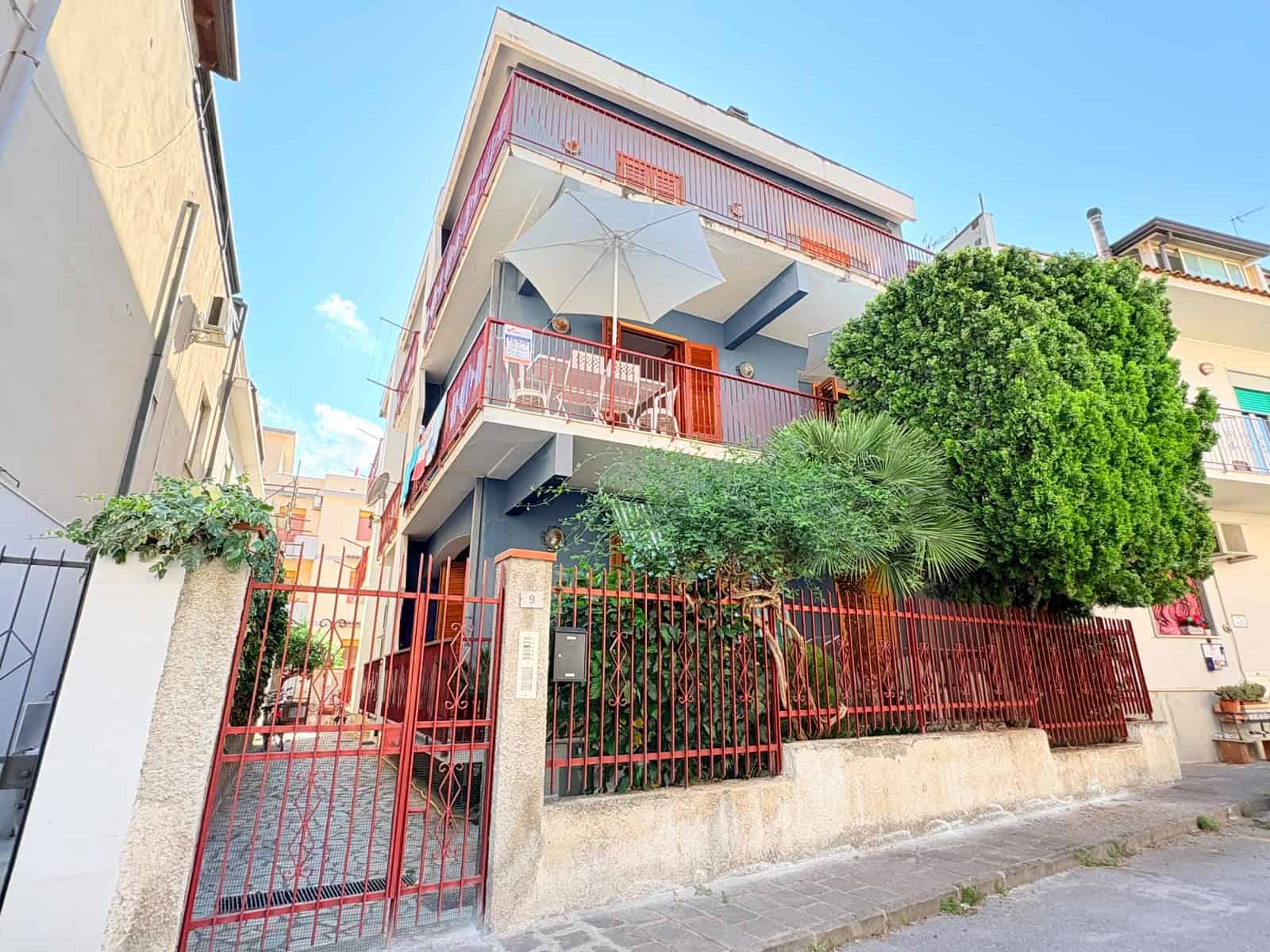 Balcone con ombrellone e piante verdi in un edificio residenziale a più piani, con recinzione rossa e accesso pedonale, in un quartiere urbano soleggiato e tranquillo, in Italia.