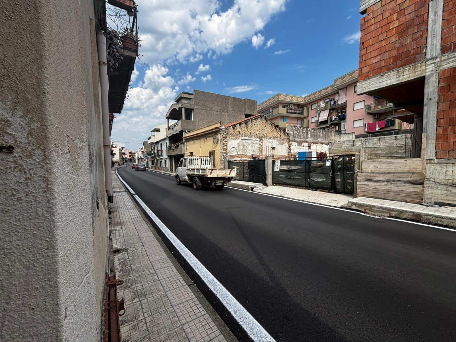 Nuova strada asfaltata in un quartiere urbano con edifici residenziali e commerciali, cielo sereno e nuvole bianche, in fase di sviluppo e rinnovamento urbano.