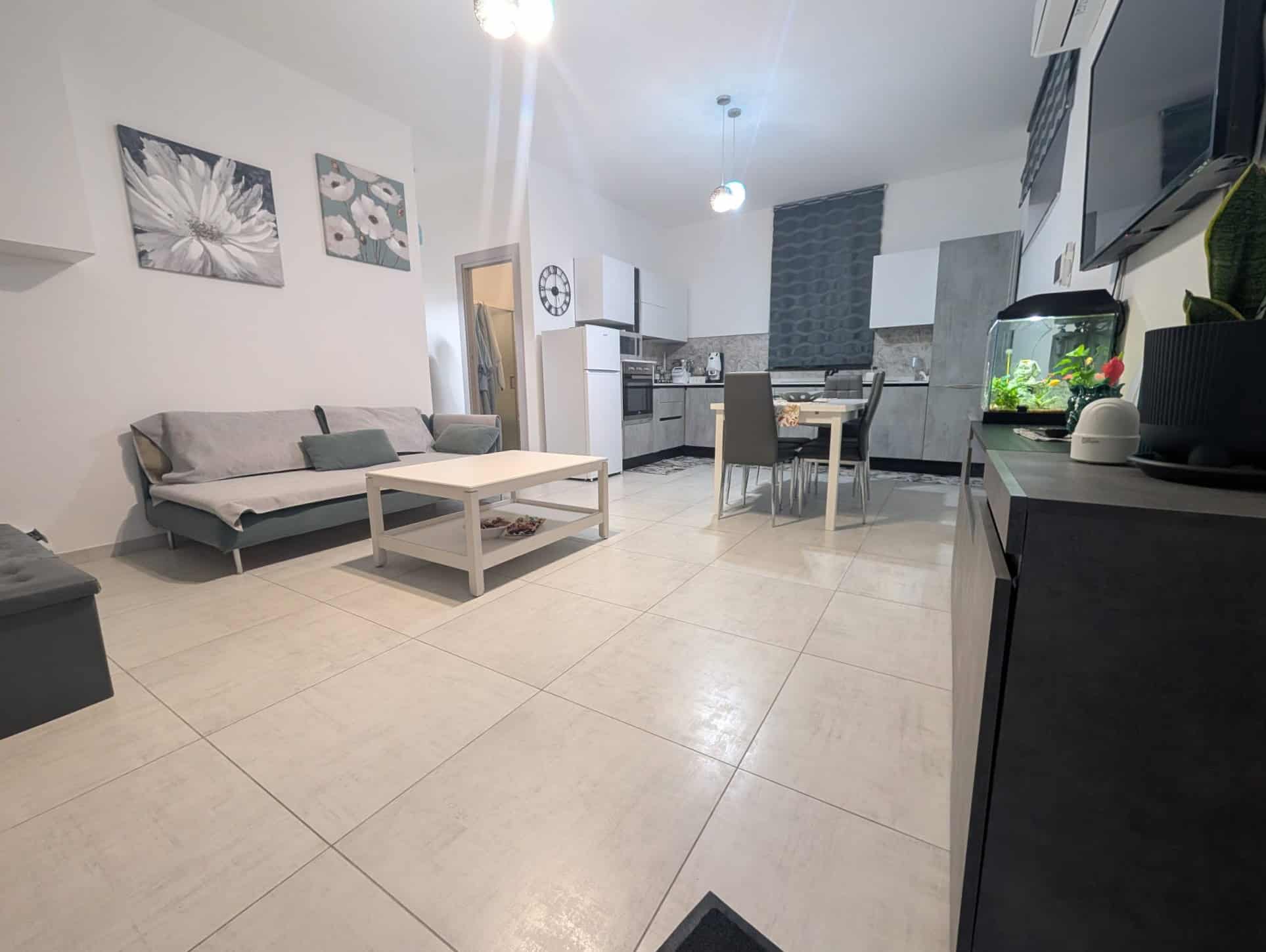Soggiorno moderno con cucina a vista, divano grigio, tavolo da pranzo e decorazioni floreali, ambiente luminoso e accogliente, ideale per vivere e rilassarsi in casa.