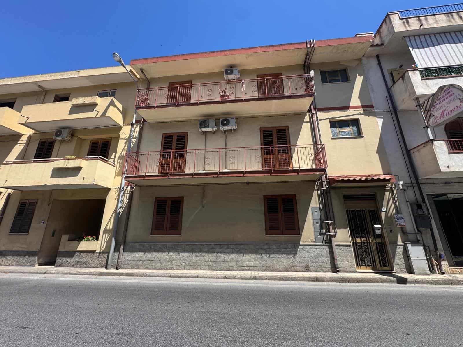 Edificio residenziale a tre piani con balconi e finestre in legno, situato lungo una strada urbana soleggiata, con aria condizionata visibile e accesso pedonale.