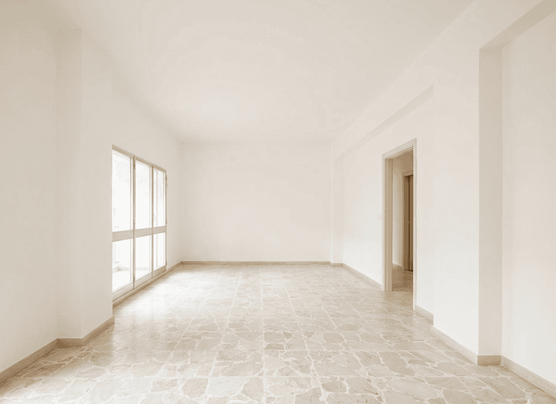 Interno di un appartamento vuoto con pareti bianche, grandi finestre e pavimento in marmo chiaro, ideale per ristrutturazione o arredamento.