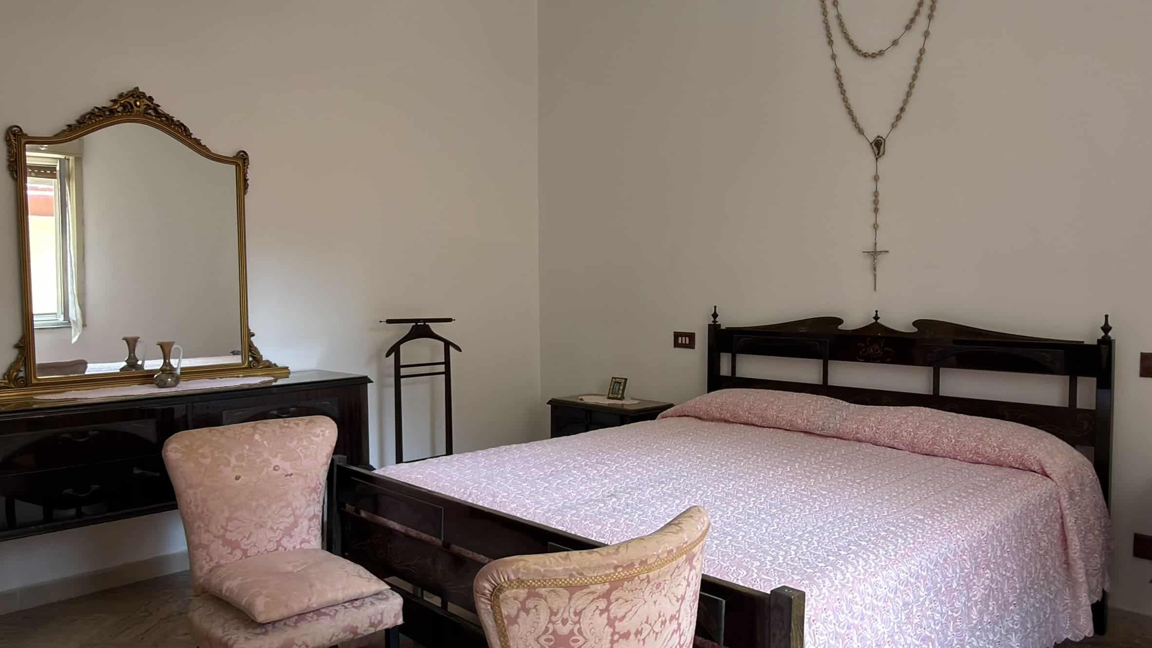 Camera da letto con letto matrimoniale in legno scuro, due sedie imbottite e un grande specchio antico, ambiente luminoso e accogliente.
