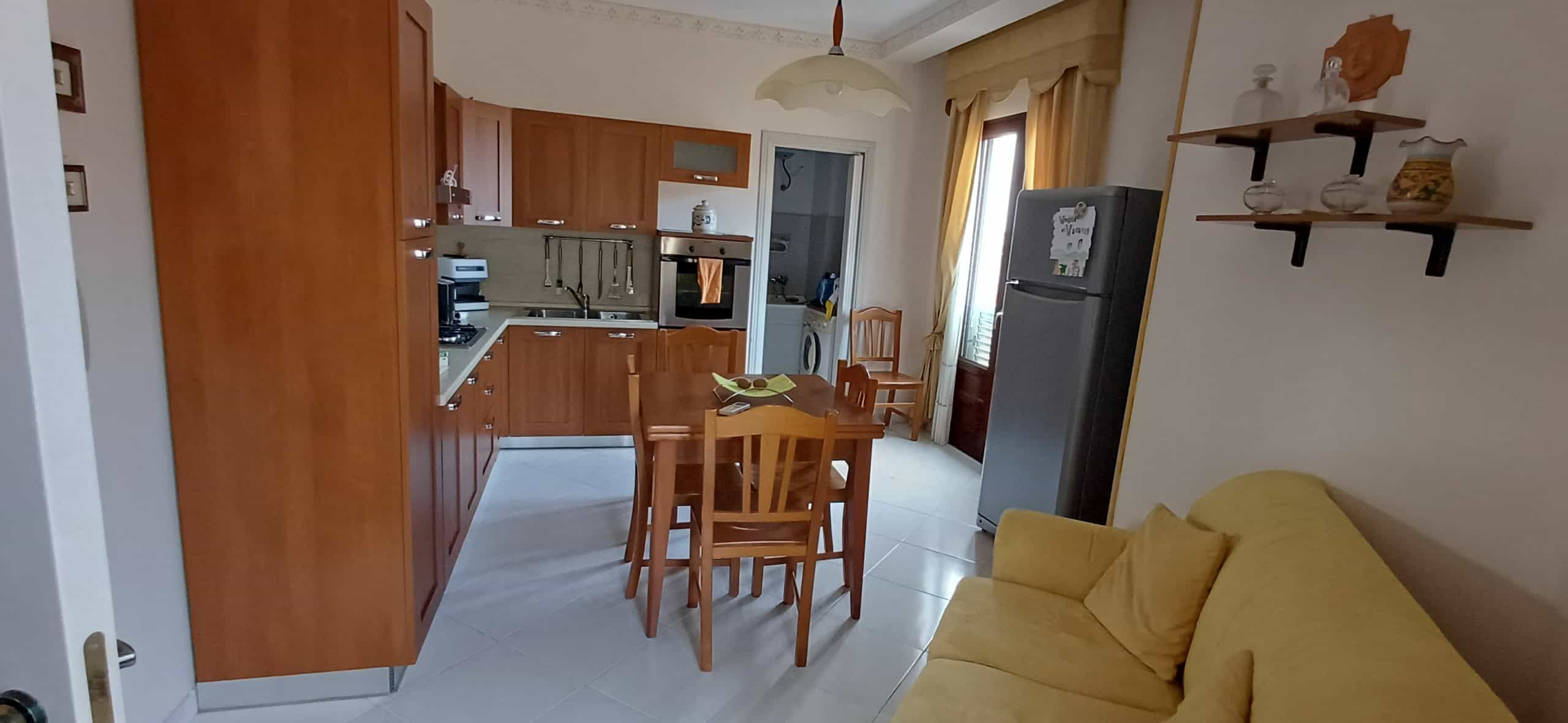 Interno di un appartamento con cucina a vista, tavolo in legno, frigorifero e divano giallo, ambiente luminoso e arredato in stile semplice e funzionale.