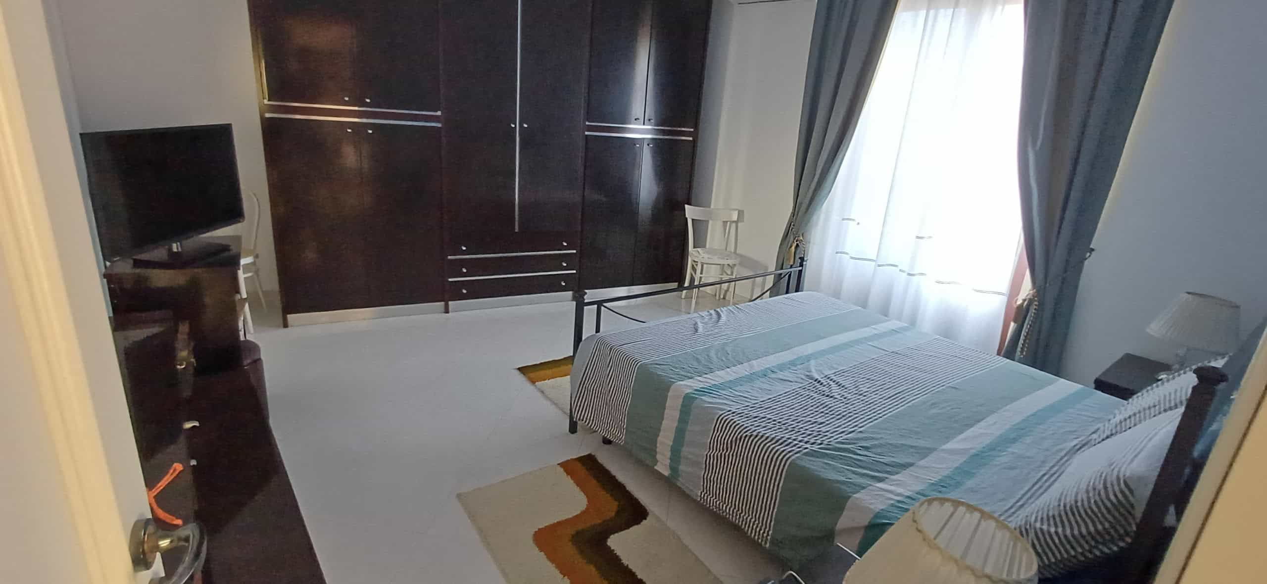 Camera da letto spaziosa con letto matrimoniale, armadio a muro, TV e finestra con tende, ambiente luminoso e accogliente.