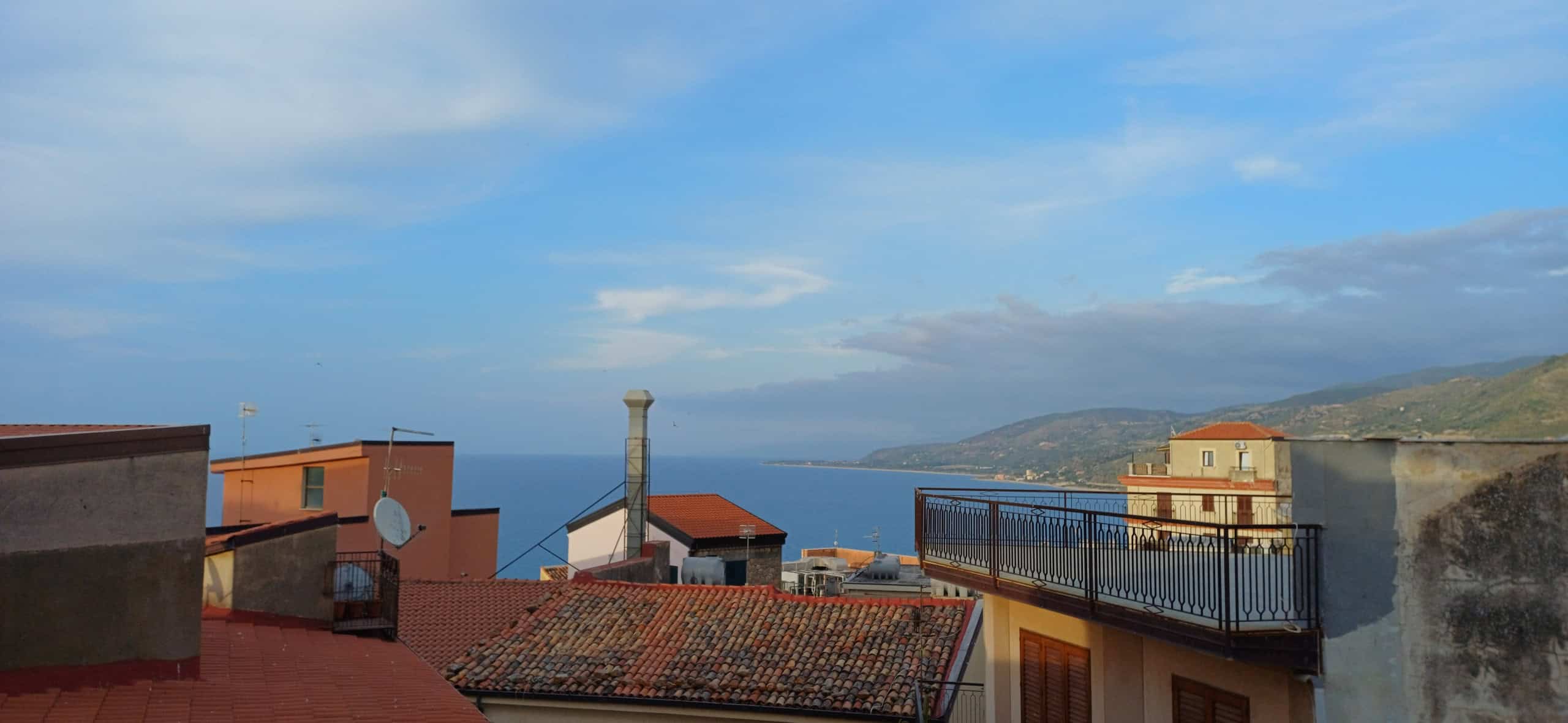 Vista panoramica di case e mare con cielo sereno e nuvole sparse, vista da un terrazzo con ringhiera in ferro, in un paesaggio costiero.