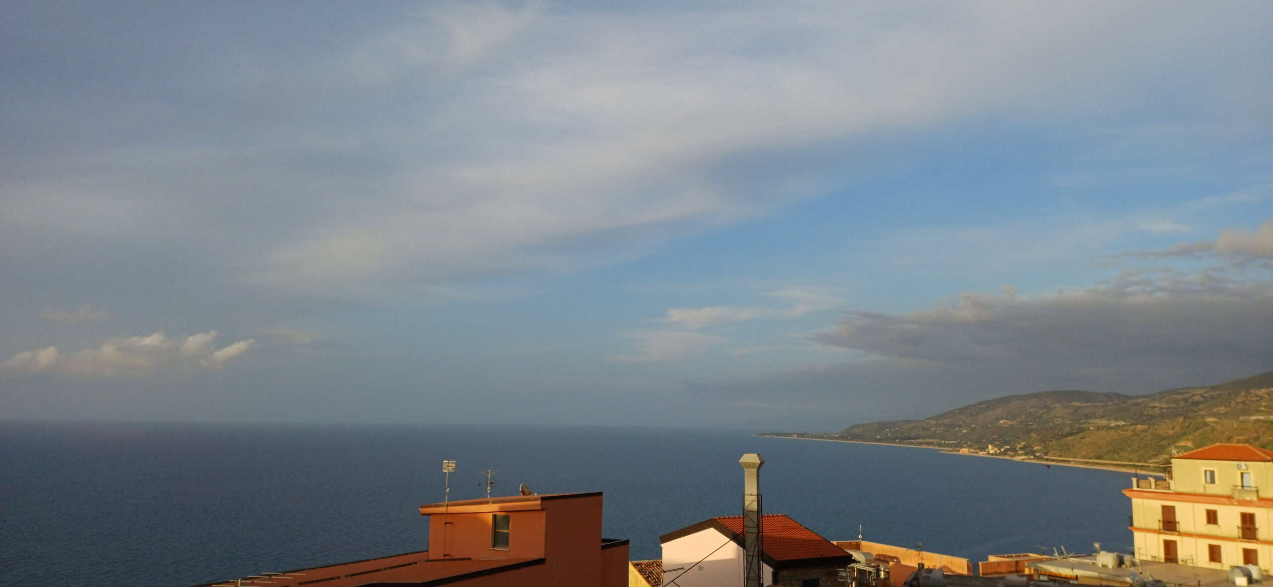 Vista panoramica sul mare e le colline con cielo nuvoloso, case sullo sfondo e paesaggio naturale.