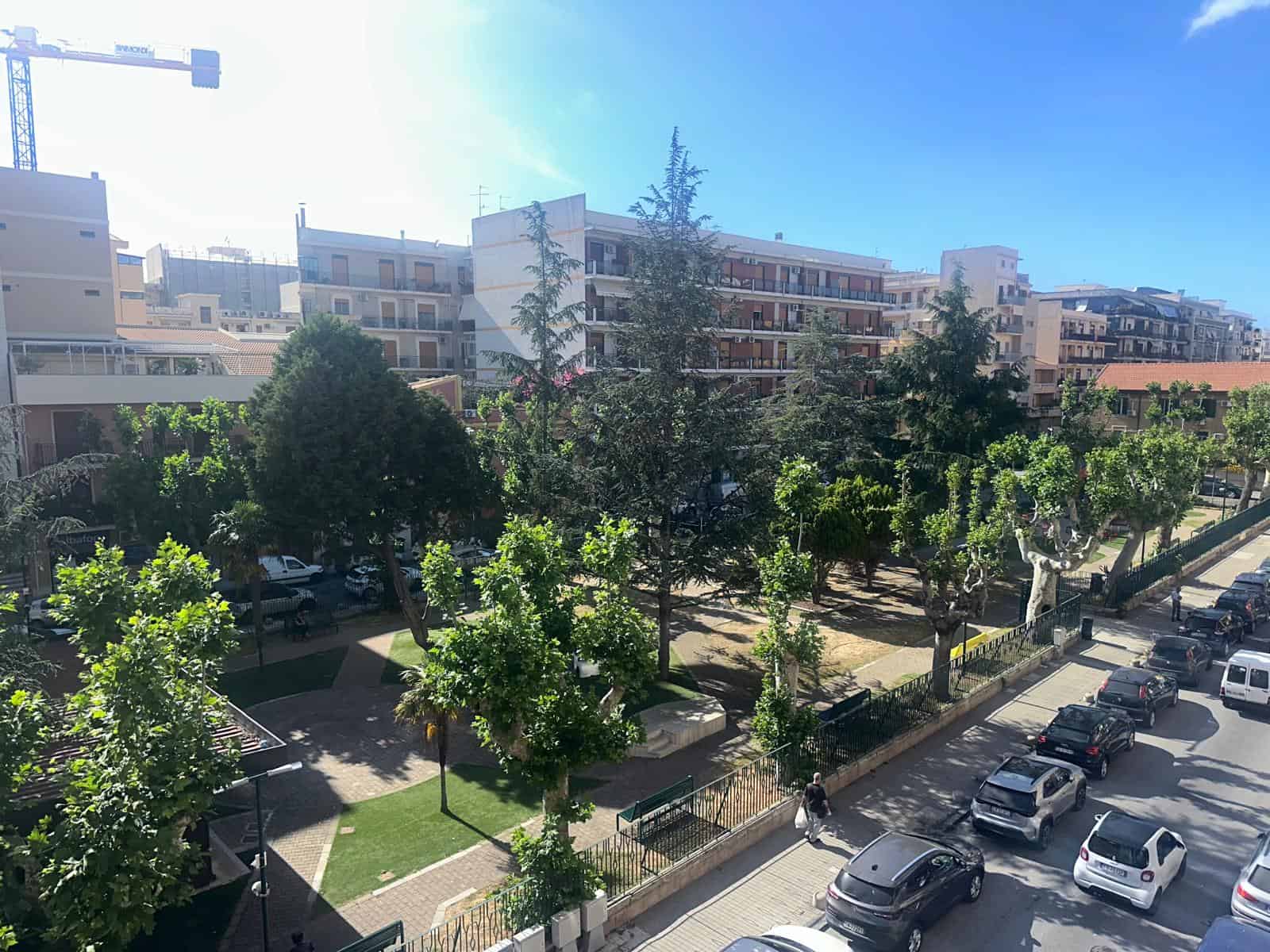 Vista panoramica di un parco urbano con alberi e panchine, situato tra edifici residenziali moderni, sotto un cielo sereno, ideale per relax e passeggiate in città.
