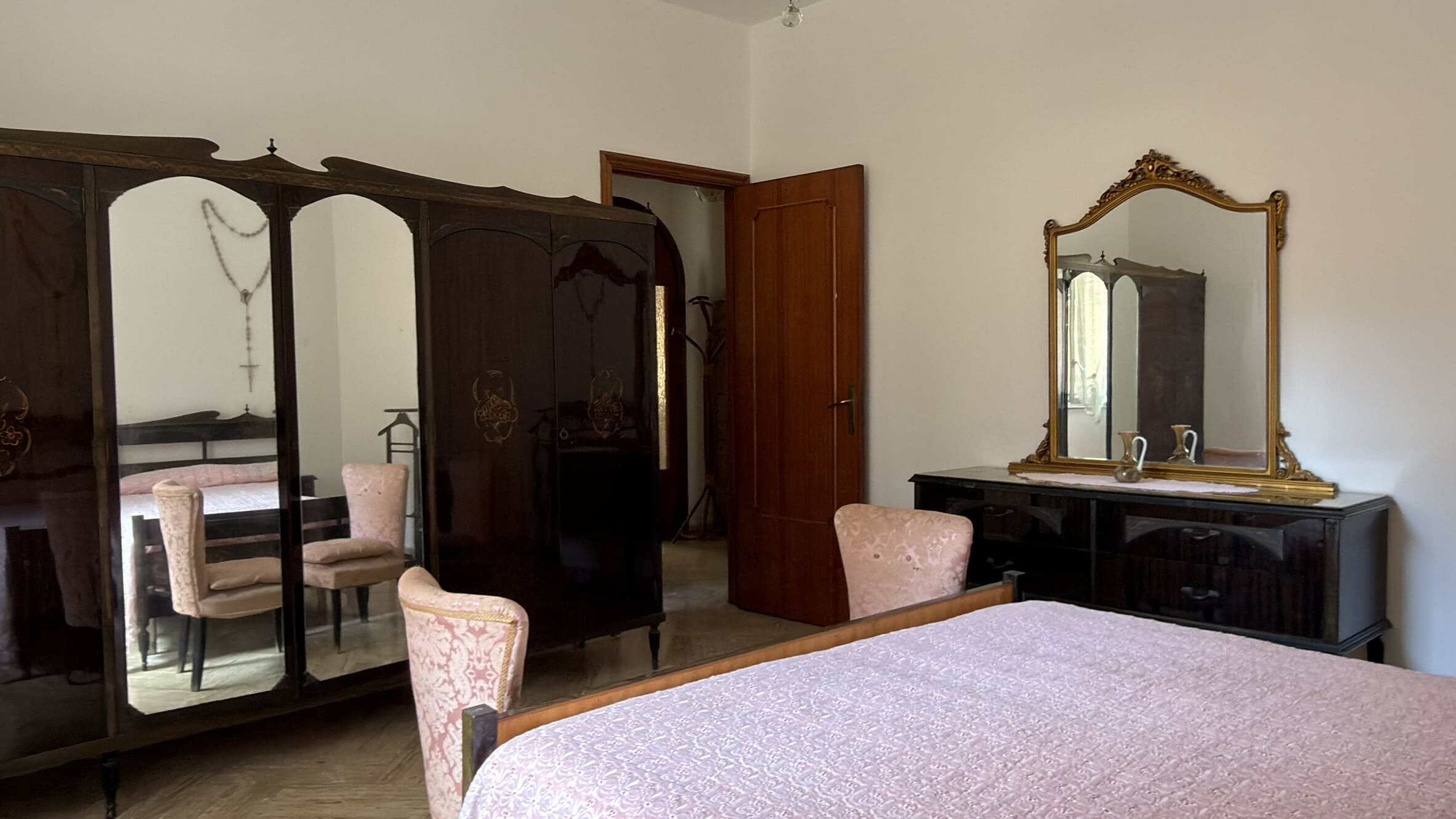 Camera che mostra una camera da letto con armadio in legno scuro, specchio e un letto con copriletto rosa, ambiente elegante e accogliente.