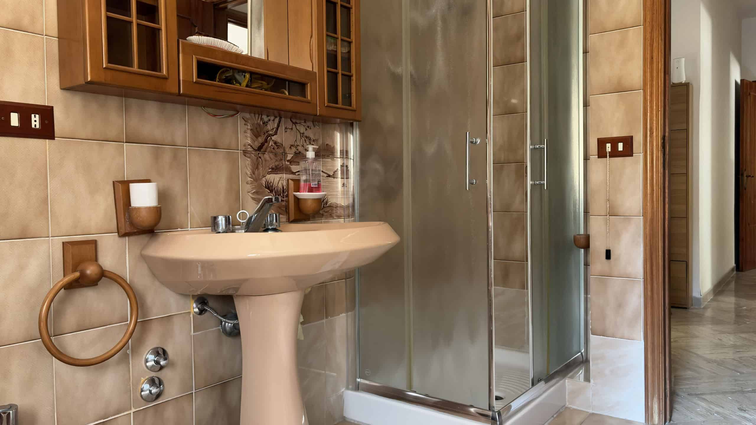 Bagno con lavabo, specchio e cabina doccia in stile classico con piastrelle beige e dettagli in legno.