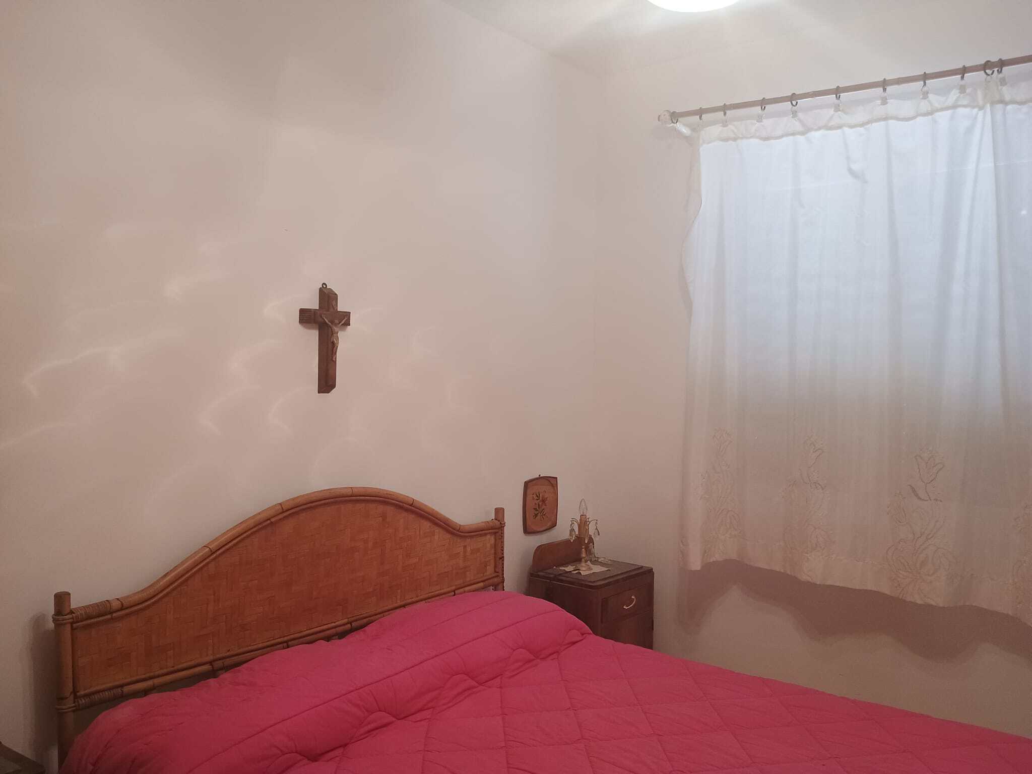 Letto matrimoniale in una stanza luminosa con decorazioni minimaliste, croce appesa sulla parete e finestra con tende trasparenti che lascia entrare la luce naturale.