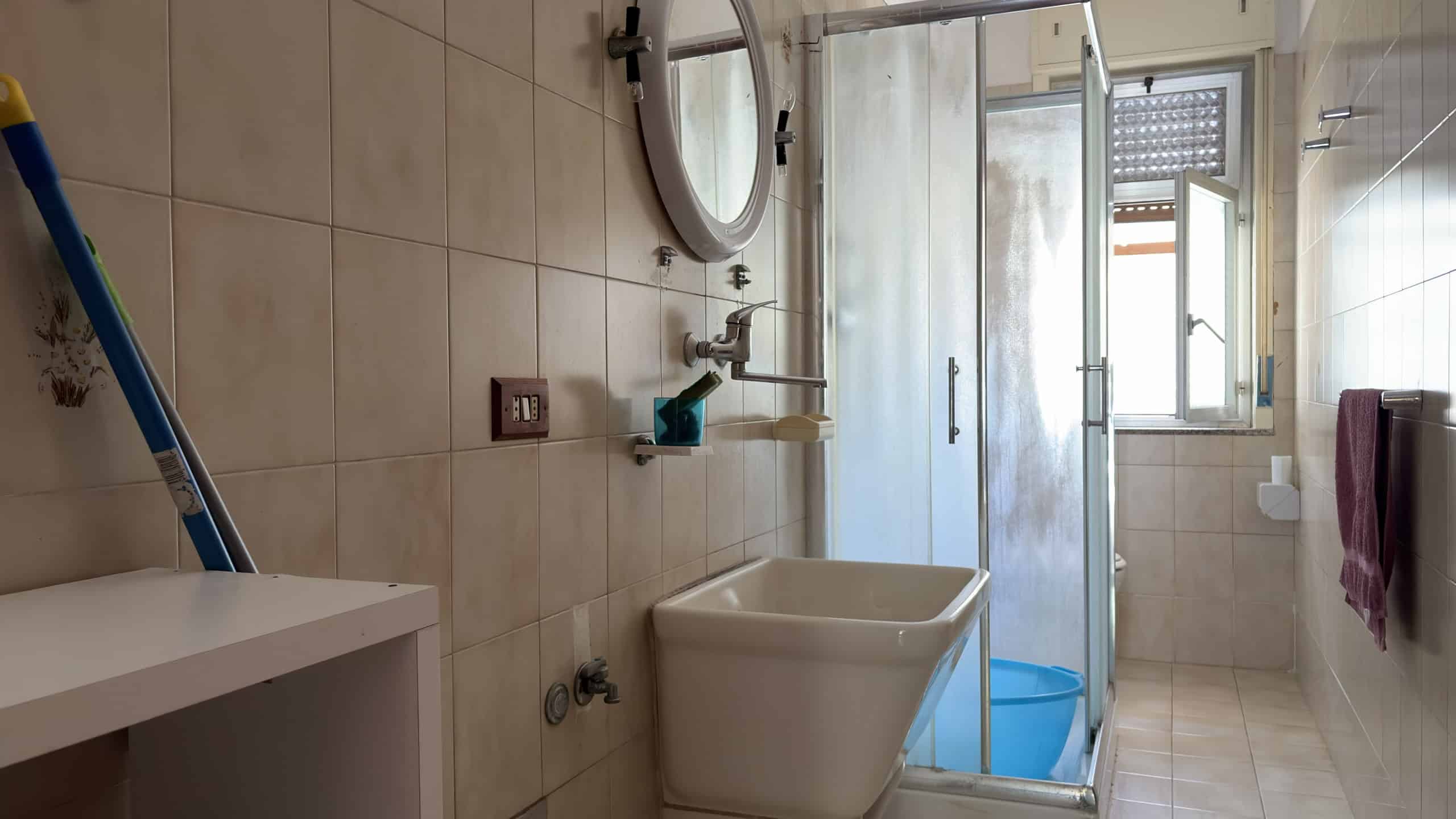 Bagno con lavabo, doccia e finestra, arredato in stile semplice e funzionale, con piastrelle beige e accessori essenziali.