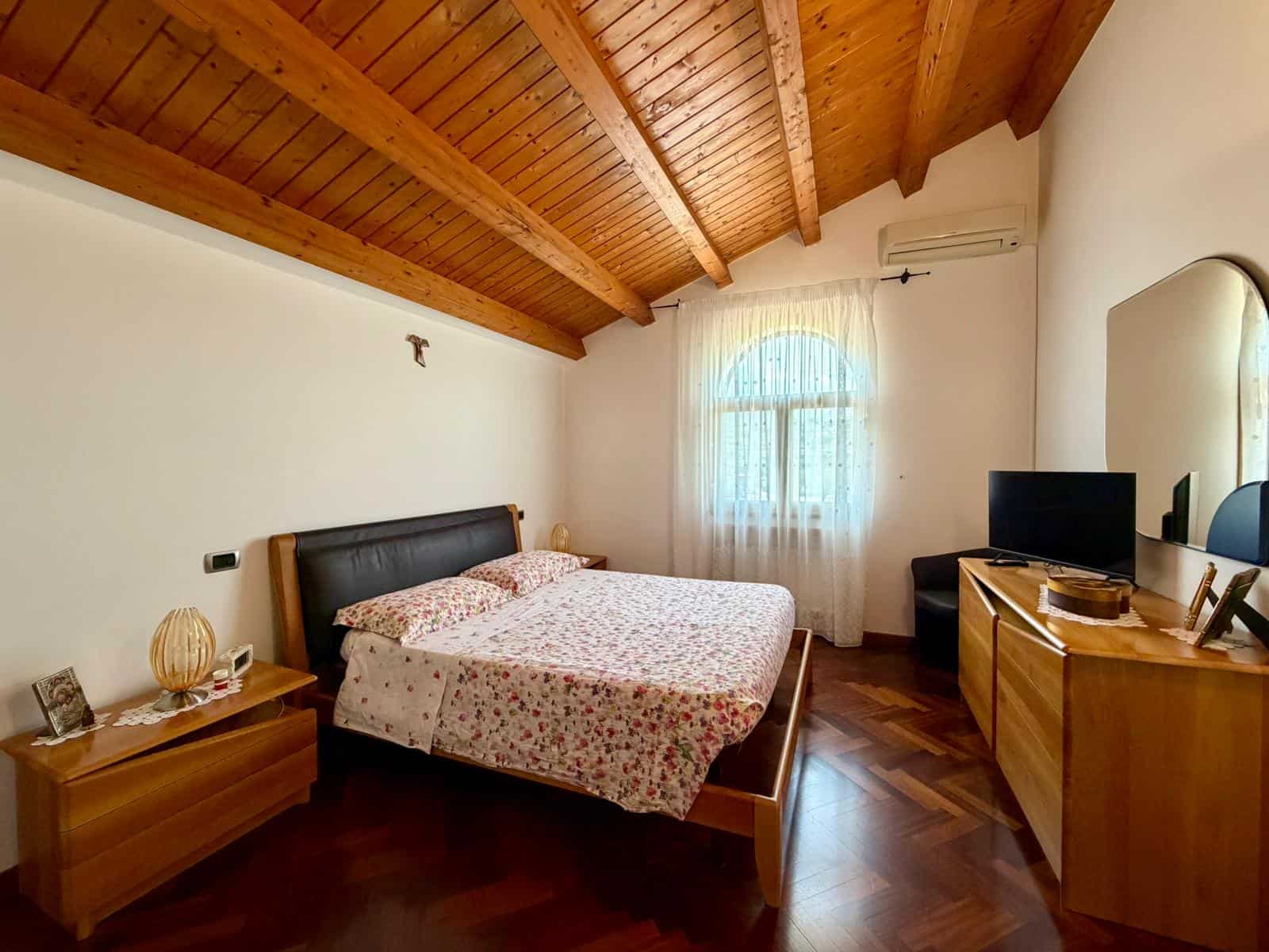 Camera da letto con soffitto in legno, letto matrimoniale, finestra con tende leggere, arredi in legno e aria condizionata.