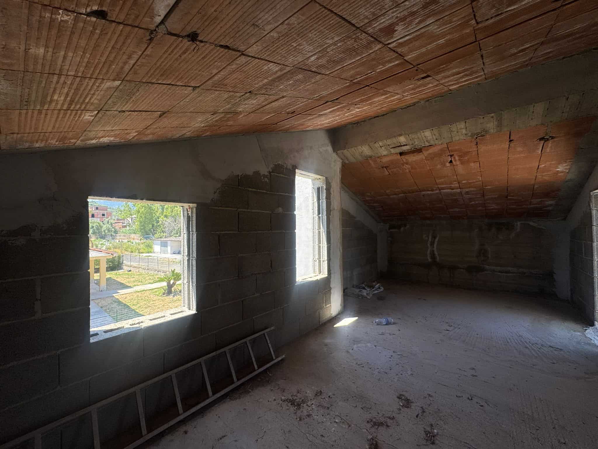 Interno di un appartamento in costruzione con pareti in cemento e soffitto in legno, luce naturale dalle finestre.