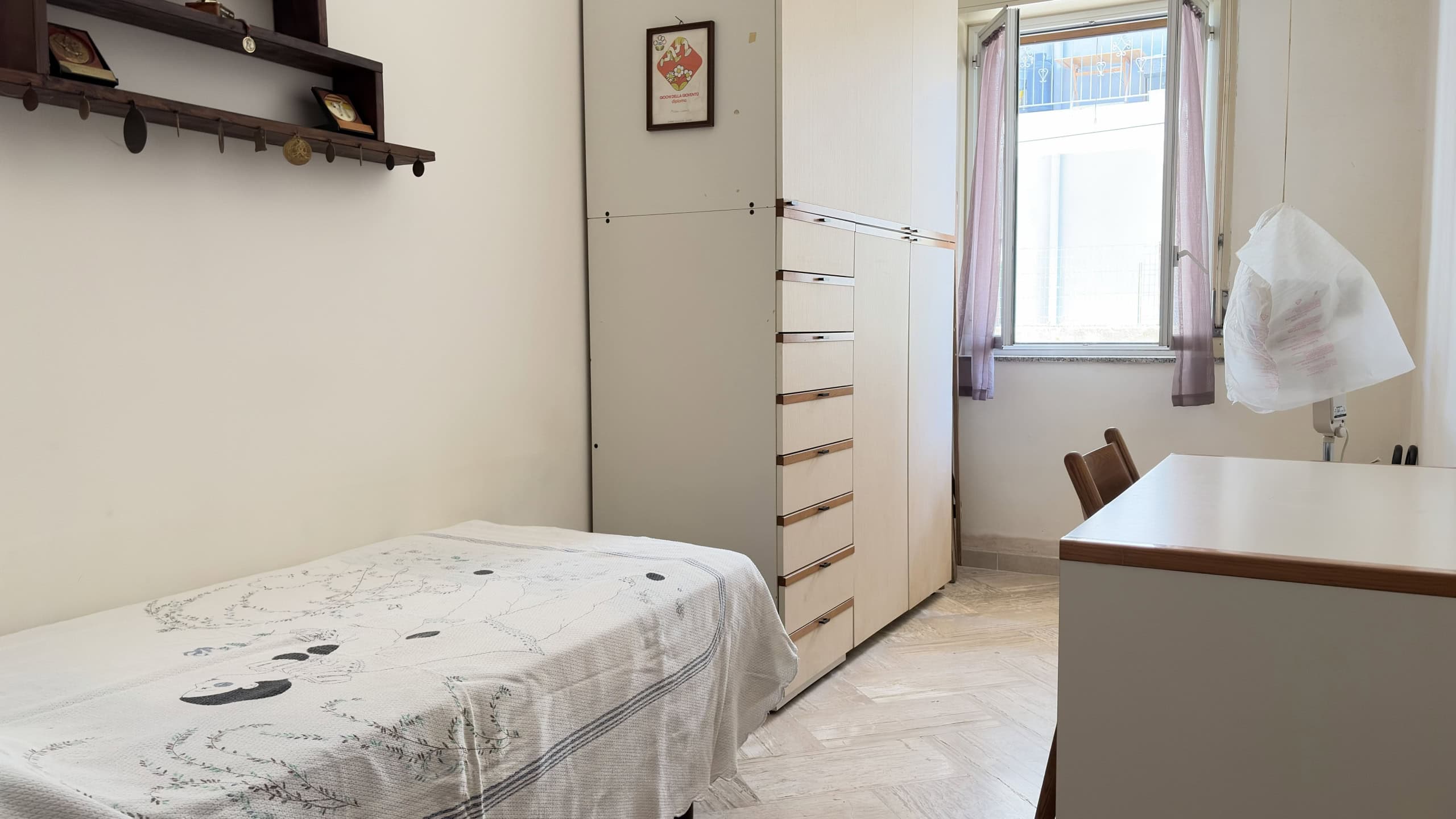 Camera da letto semplice con letto singolo, armadio e scrivania, illuminata da una finestra con tende lilla.