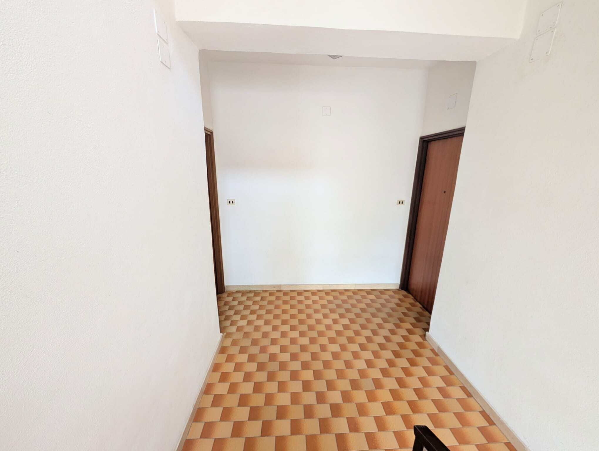 Ingresso di un appartamento con pareti bianche e pavimento in piastrelle a scacchi marroni e beige, con due porte in legno scuro.