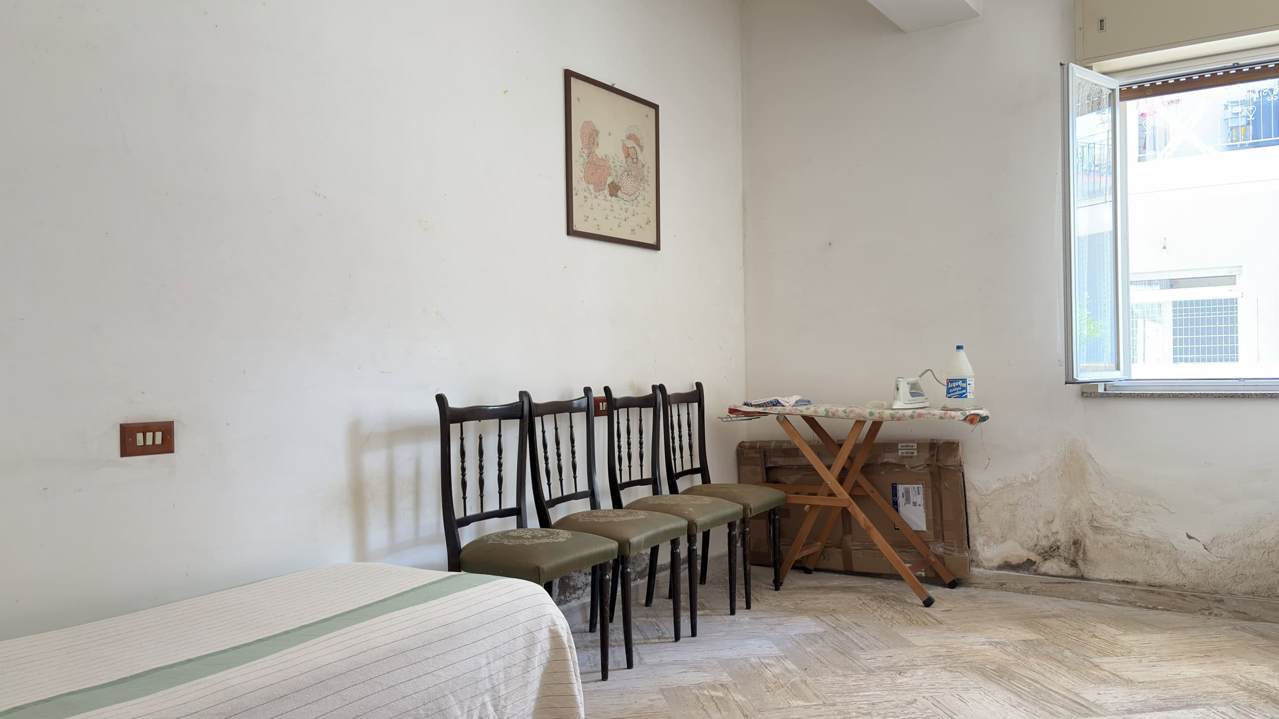 Interno di una stanza con pareti bianche, quattro sedie di legno e un letto parzialmente visibile, finestra aperta con vista su edifici.