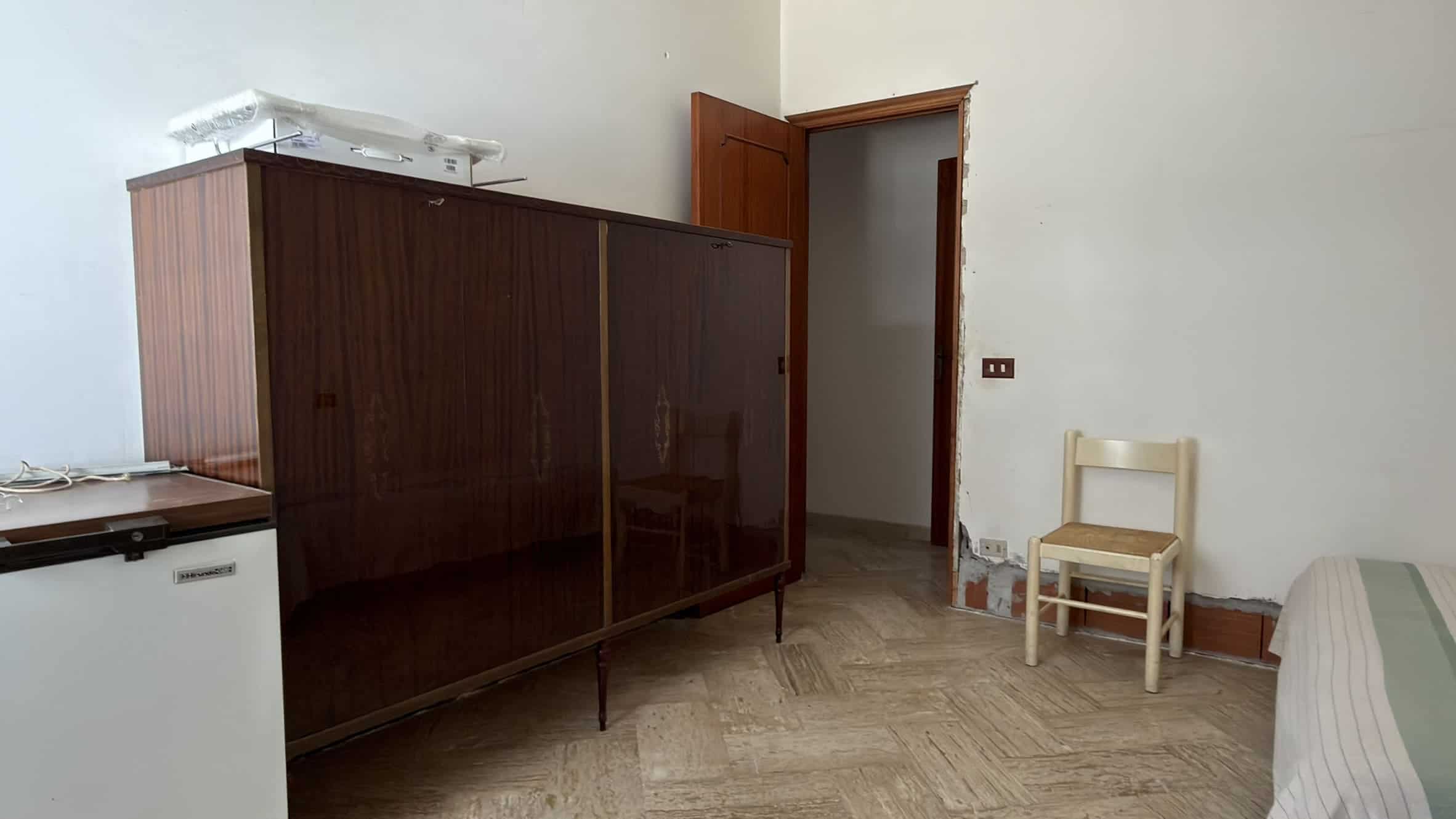 Interno di una stanza con armadio in legno scuro, sedia bianca e pareti bianche, pavimento in piastrelle beige.