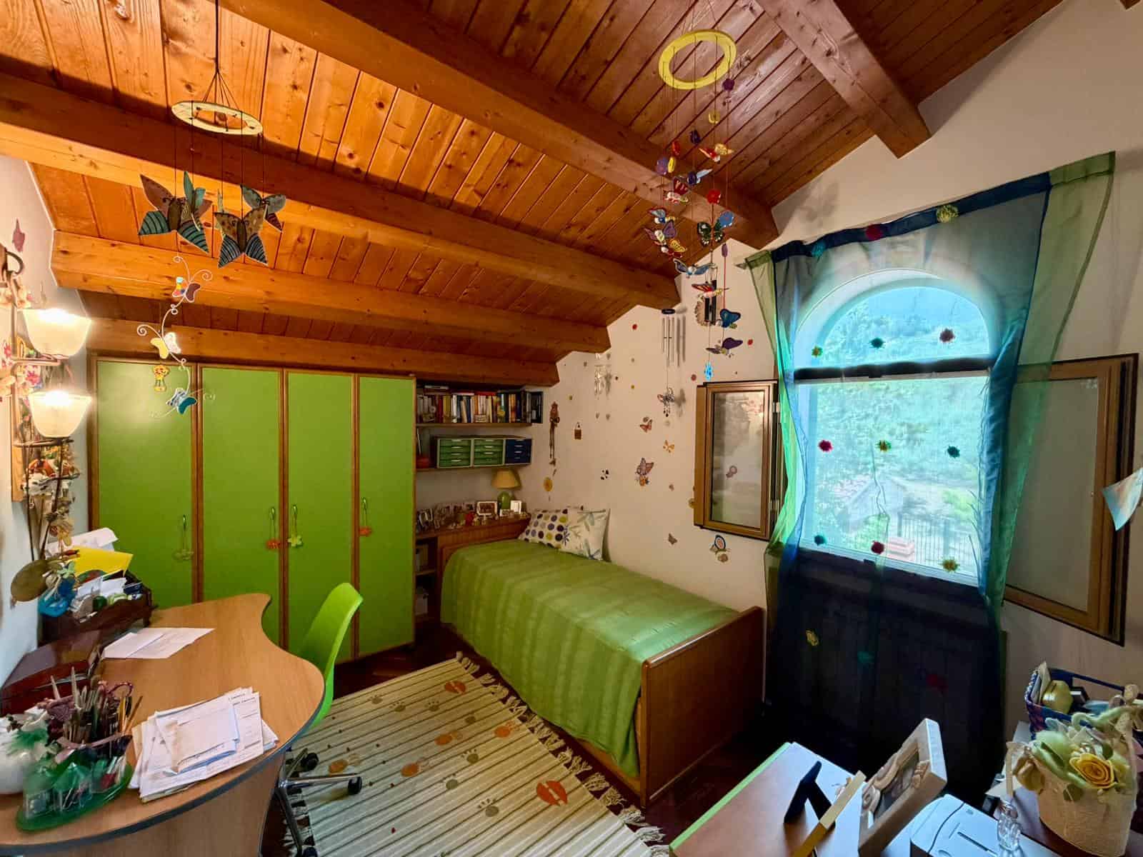 Camera da letto in stile rustico con soffitto in legno, armadio verde, finestra con tende azzurre e decorazioni colorate.