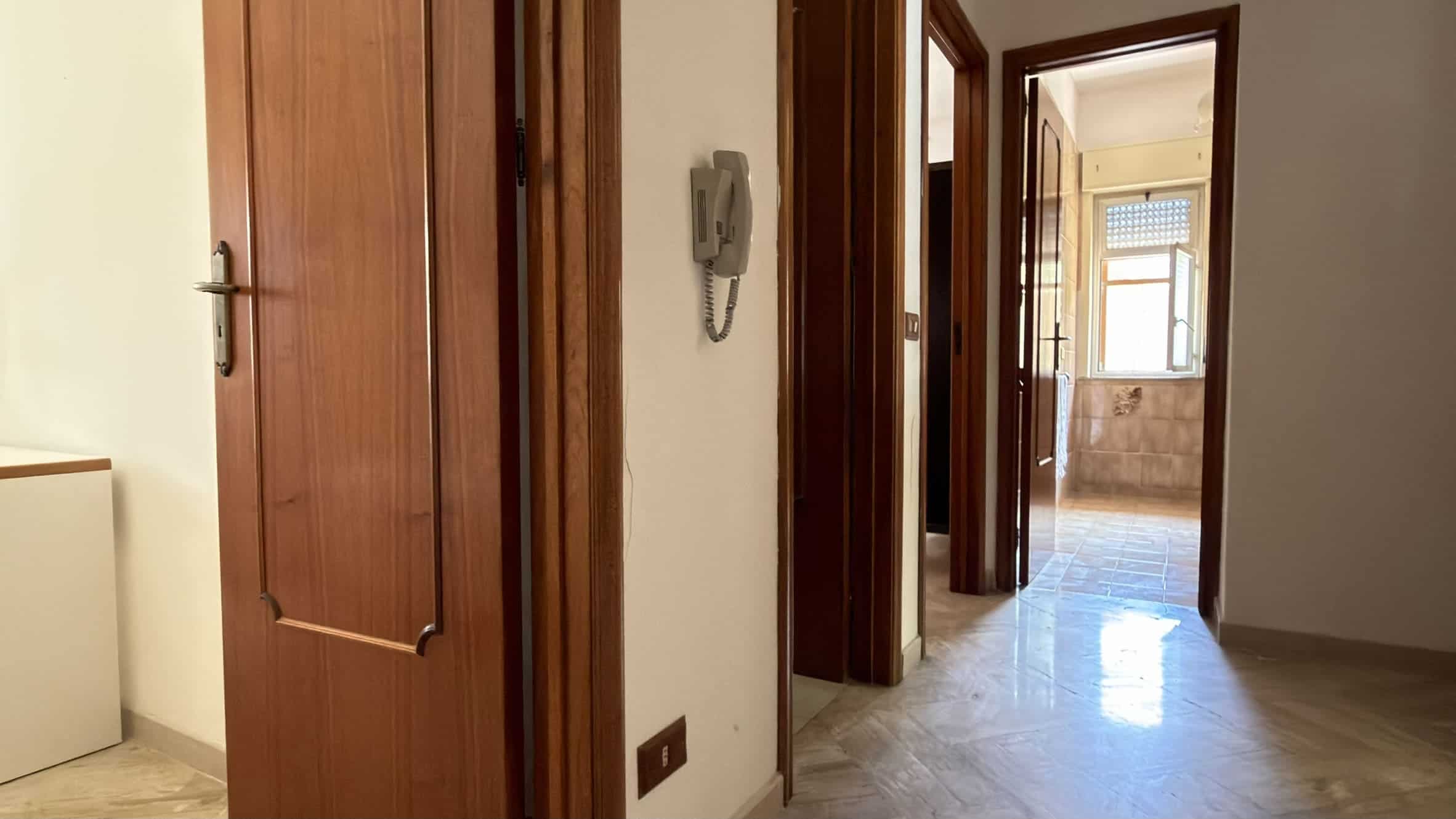 Interno di un appartamento con porte in legno e pavimento in marmo, vista verso una stanza luminosa con finestra.